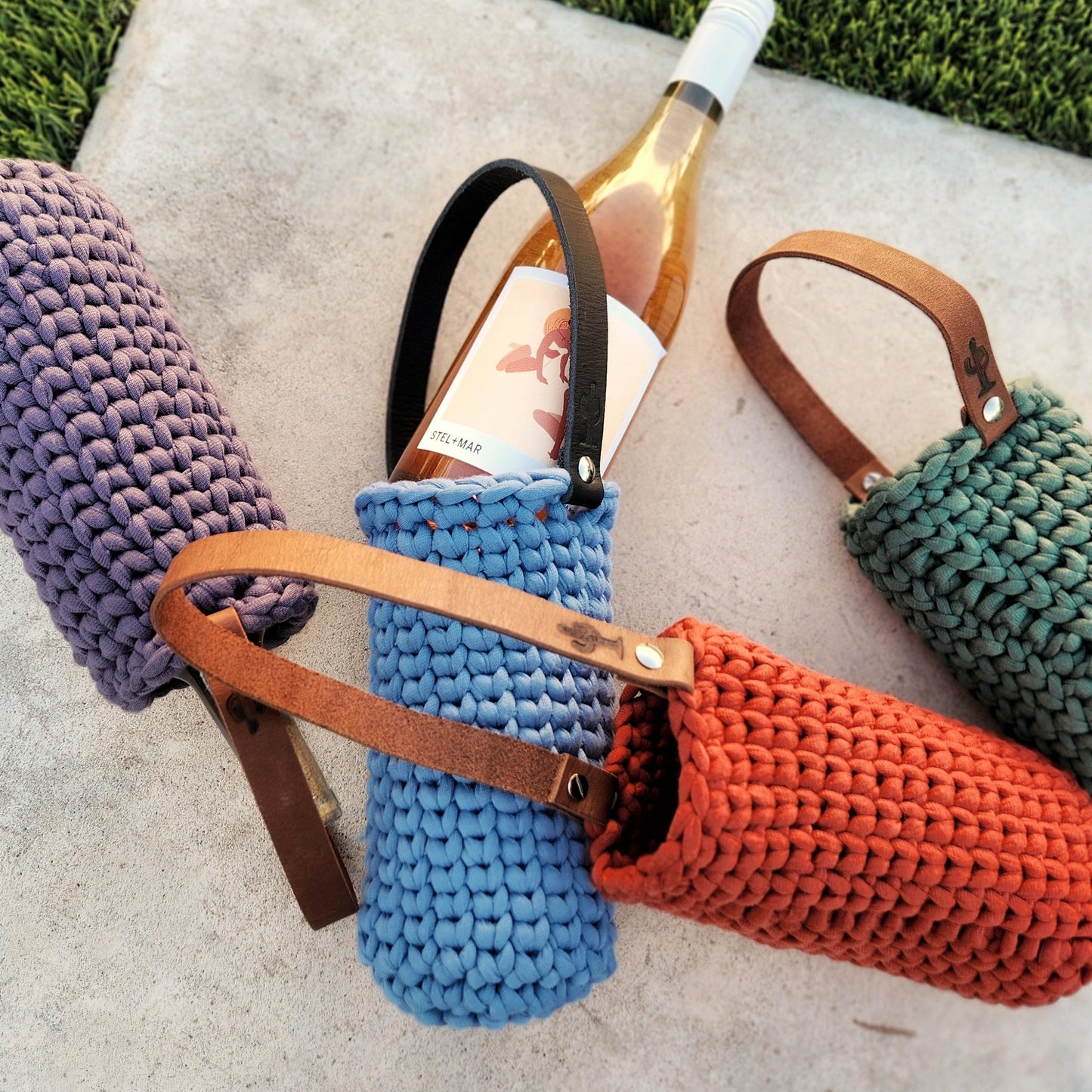 A collection of hand-crocheted wine bottle carriers in rich colors—purple, blue, red-orange, and green—each featuring a sturdy leather handle with cactus branding. A bottle of rosé peeks out of the blue holder, showcasing the carrier’s snug and stylish fit. The vibrant yarn textures and natural leather contrast beautifully against the concrete and grass background, making these eco-friendly carriers both functional and gift-ready.