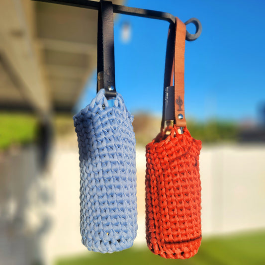 Two hand-crocheted wine bottle carriers hang from a metal rod outdoors—one in denim blue and the other in pumpkin orange. Each has a sturdy leather strap with a stamped cactus design and holds a wine bottle inside.