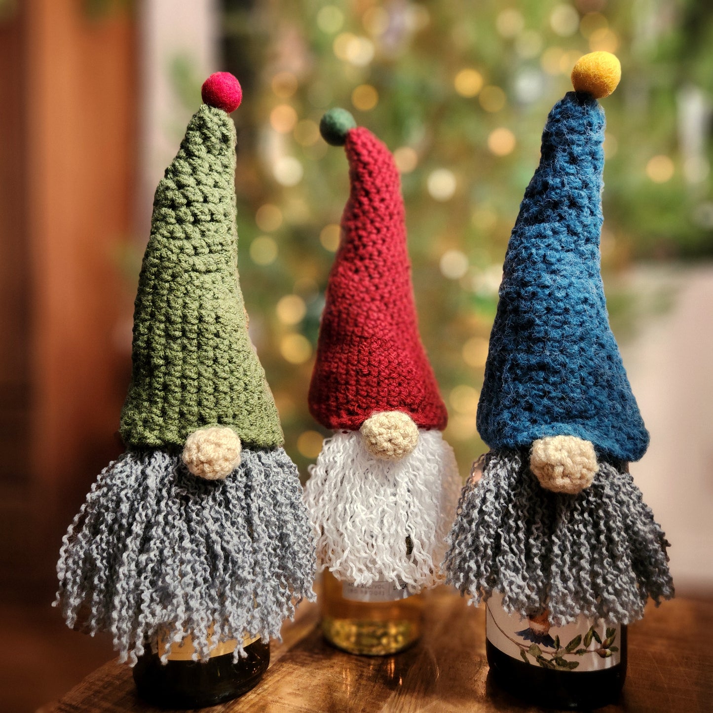 Three hand-crocheted holiday gnome bottle toppers displayed on wine bottles. Each gnome features a tall, pointed hat—green with a red pom-pom, red with a green pom-pom, and blue with a yellow pom-pom—and long, curly yarn beards. Perfect as festive wine bottle decorations or handmade holiday gifts, set against a softly lit Christmas tree background.