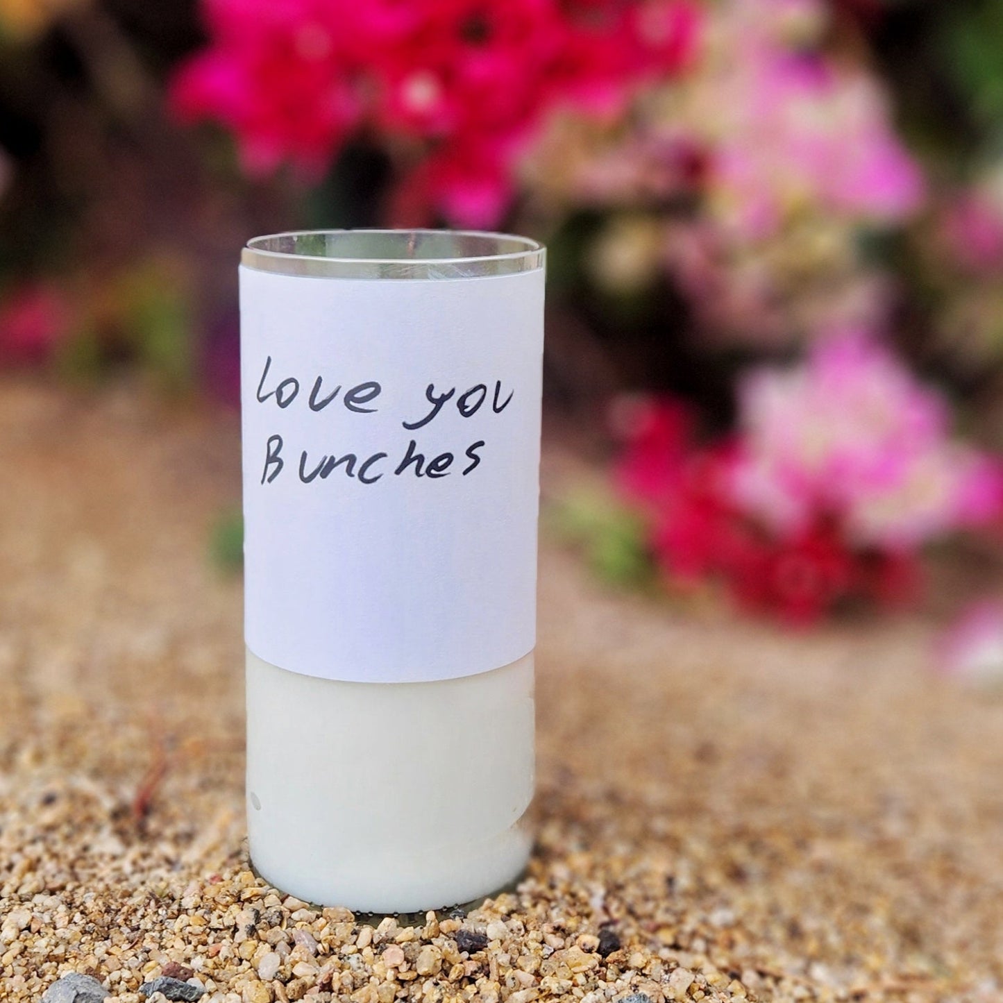 A hand-poured coconut soy wine bottle candle crafted from a recycled clear glass bottle. The minimalist label features a handwritten-style message that reads, Love You Bunches, adding a personal and heartfelt touch. This eco-friendly wooden wick candle has a smooth, polished glass rim, highlighting expert craftsmanship. Placed on a natural desert ground with vibrant pink bougainvillea flowers in the background, this sustainable wine bottle candle makes a thoughtful gift for wine lovers and eco-conscious sho