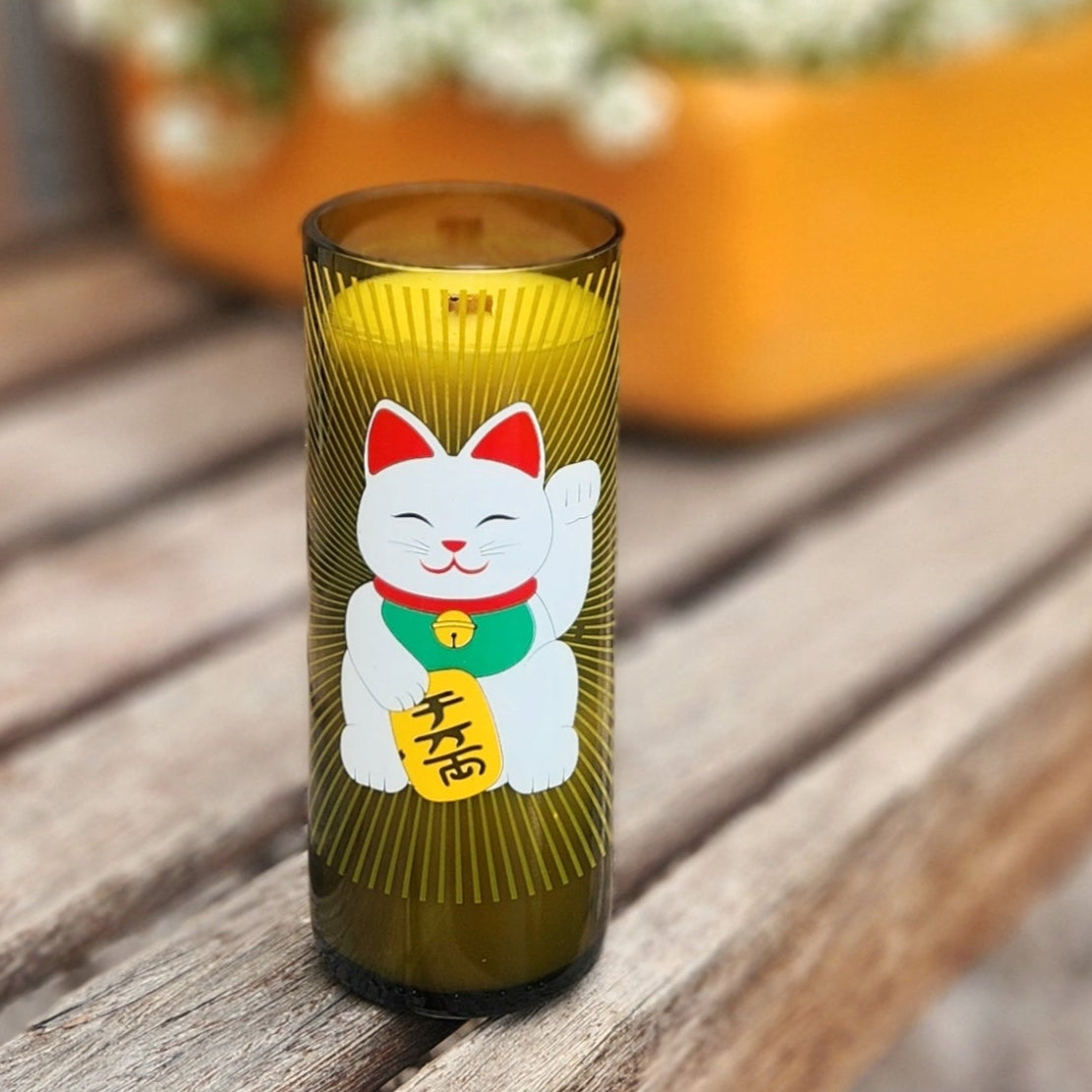 A handcrafted candle made from a recycled green wine bottle, featuring a vibrant Lucky Cat (Maneki-Neko) design. The label showcases a cheerful white cat with a raised paw, a green collar, and a gold coin with Japanese characters, set against a radiant golden sunburst pattern. The candle sits on a wooden bench outdoors, with a blurred background of blooming white flowers and a bright yellow planter, creating a warm and inviting atmosphere. The smooth glass rim and golden wax enhance its artisanal charm.