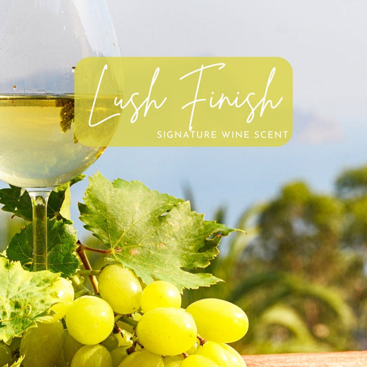 A wine glass filled with chilled white wine rests next to a bunch of fresh green grapes and leafy vines on a sunny deck. The text “Lush Finish - Signature Wine Scent” overlays the image, evoking a tropical white wine-inspired candle fragrance.