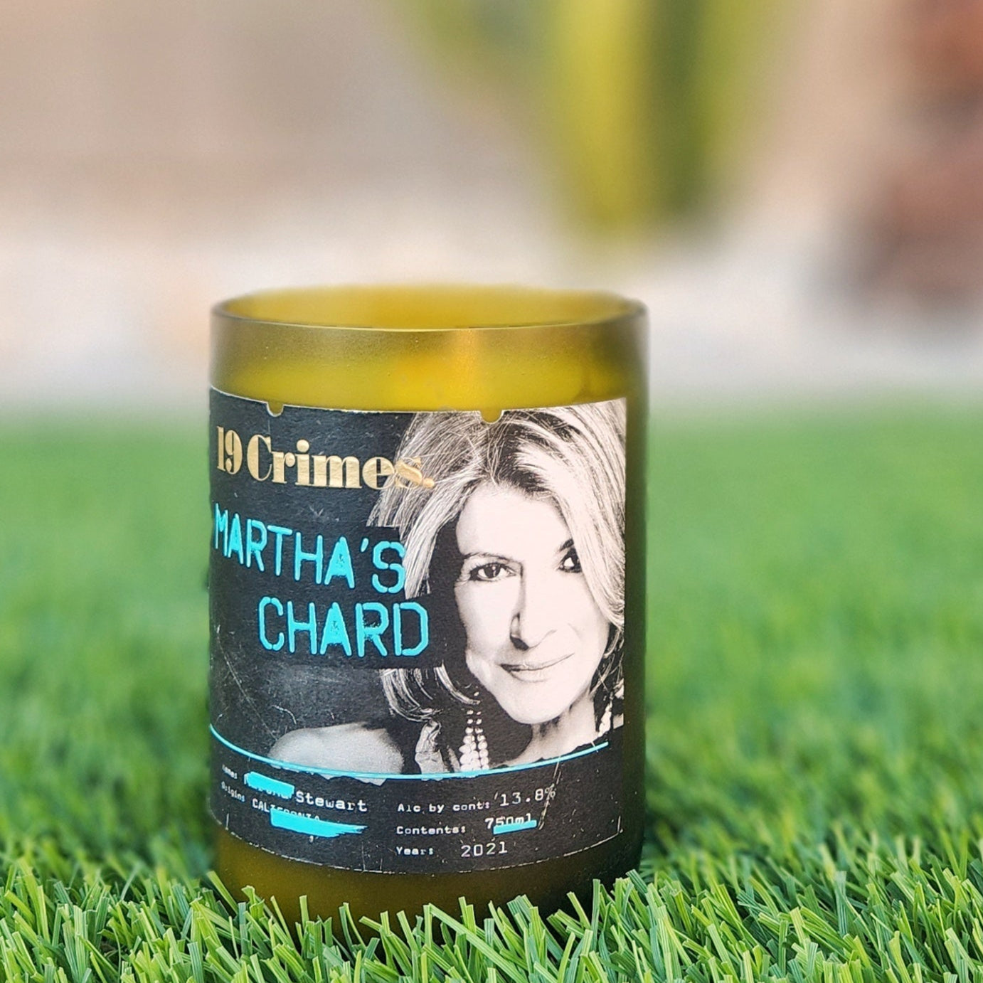 A hand-poured candle crafted from coconut-soy wax, housed in a recycled green wine bottle with a smooth, frosted rim. The label features a design from "19 Crimes Martha's Chard," showcasing a portrait of Martha Stewart in bold, modern typography. The candle is placed on a lush patch of artificial grass, with a blurred background of a cactus and a decorative sculpture, adding a stylish and eco-friendly vibe to the scene. Perfect for upcycled wine bottle decor.