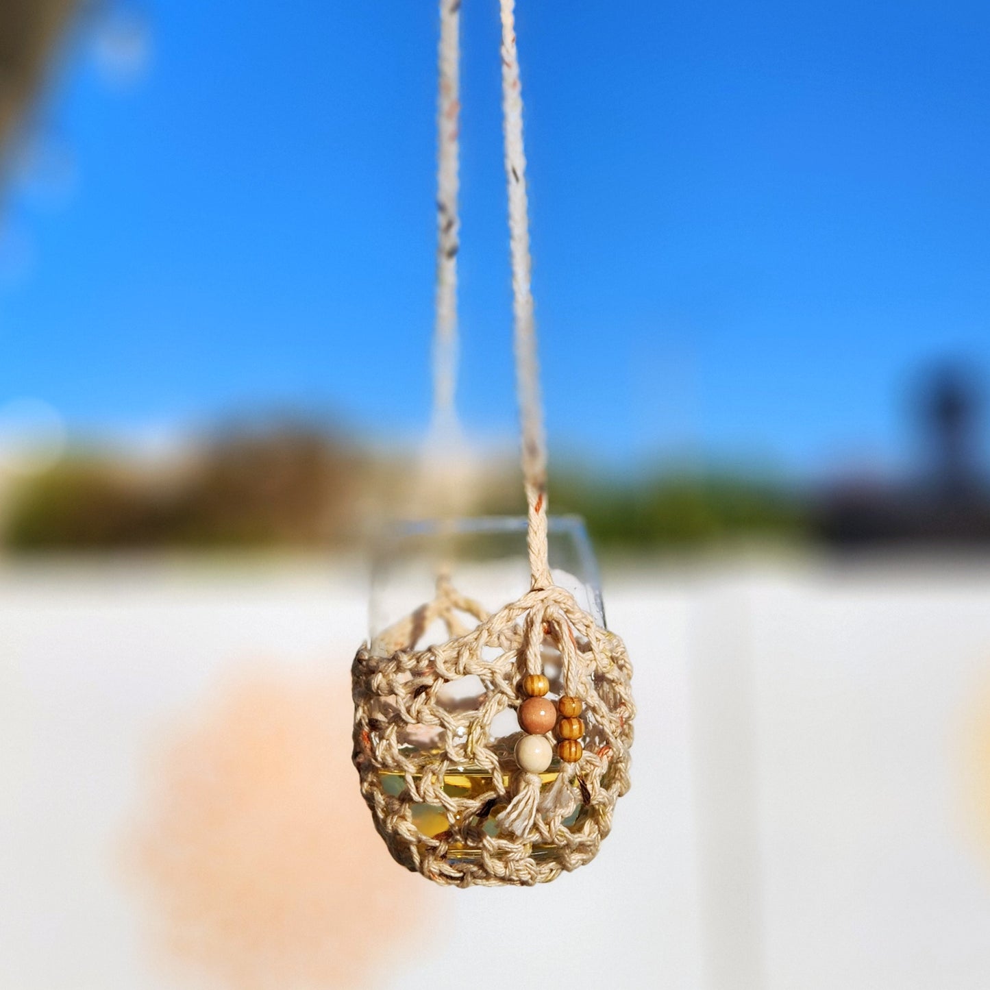 Handmade macramé wine glass holder featuring neutral-toned yarn and natural wood beads, suspending a clear wine glass against a bright blue sky. This boho-style wine glass lanyard adds a charming, hands-free touch to outdoor sipping.