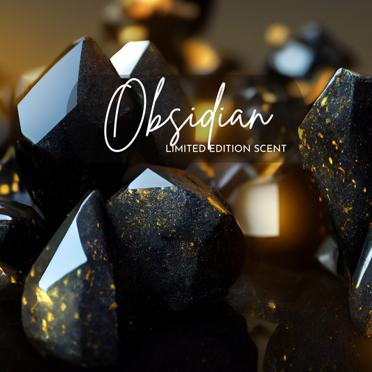 A close-up of glossy black obsidian crystals with golden flecks, arranged on a dark reflective surface. The lighting adds a warm glow that highlights the luxurious, dramatic tone of this limited edition Obsidian scent image. Text overlay reads “Obsidian – Limited Edition Scent” in elegant script.