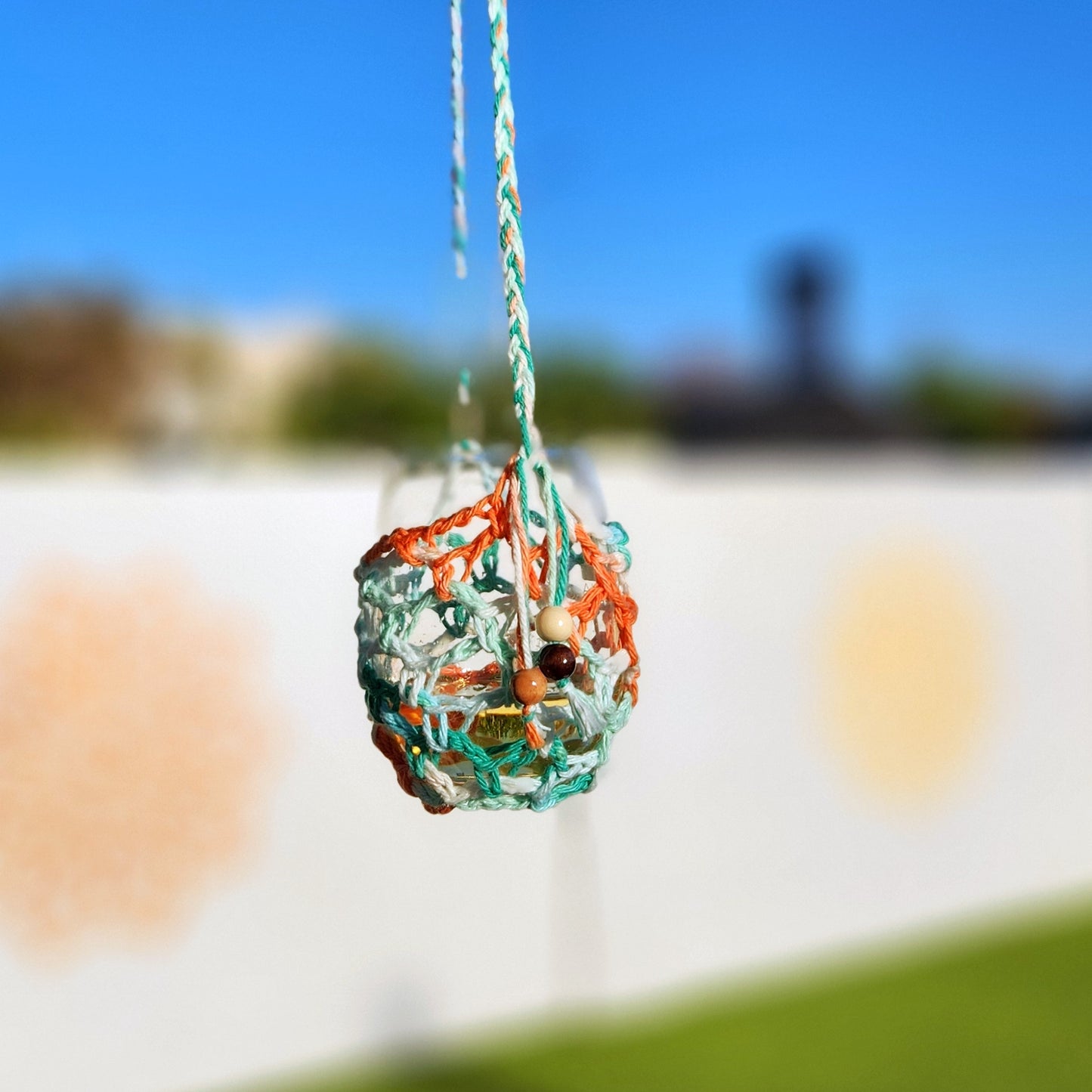 Close-up of a hanging wine glass in a summery orange and mint-colored crocheted holder with braided strap and small beaded tassels.