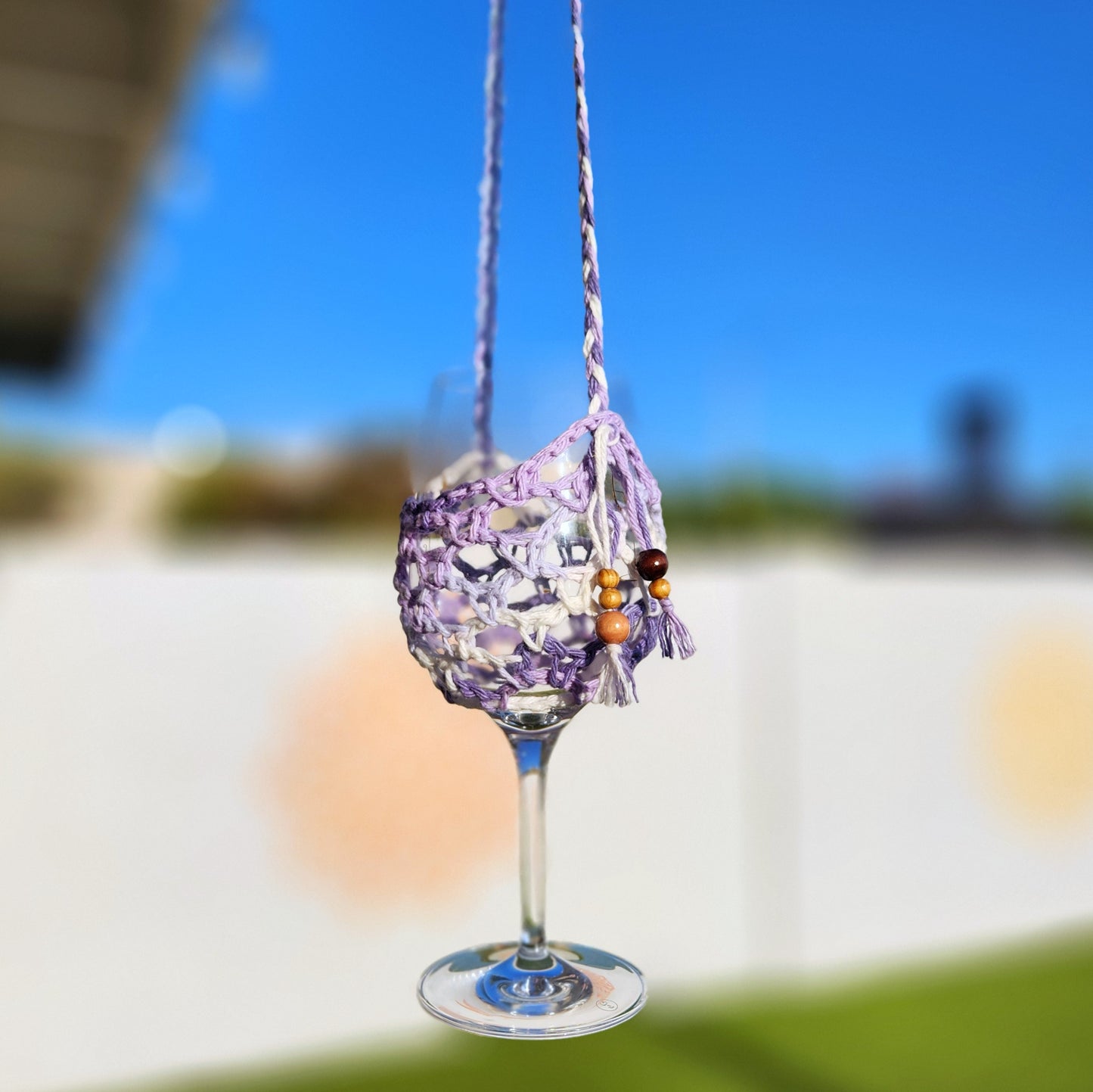 A close-up of a purple and white crochet wine glass lanyard with beaded accents, hanging outdoors against a blurred sunny backyard scene. The glass is supported by a netted yarn holder suspended by a braided strap.