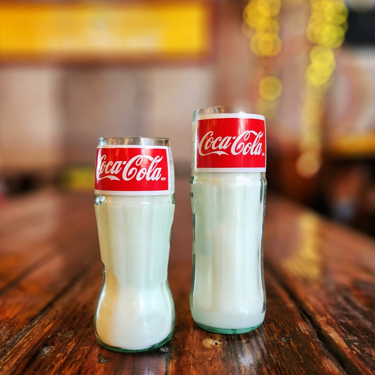 Two upcycled Coca-Cola bottle candles displayed side by side on a wooden surface, featuring eco-friendly coconut soy wax and red branded labels.