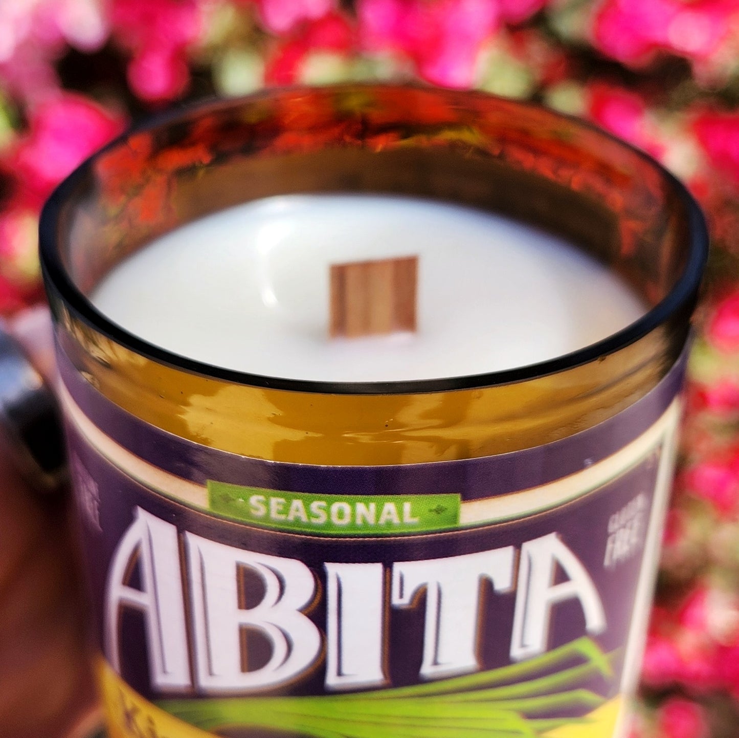 A close-up of a sustainable amber glass candle, hand poured with creamy white coconut soy wax and a crackling wooden wick. The smooth, polished rim catches the sunlight, with the festive King Cake-themed label wrapping the front.
