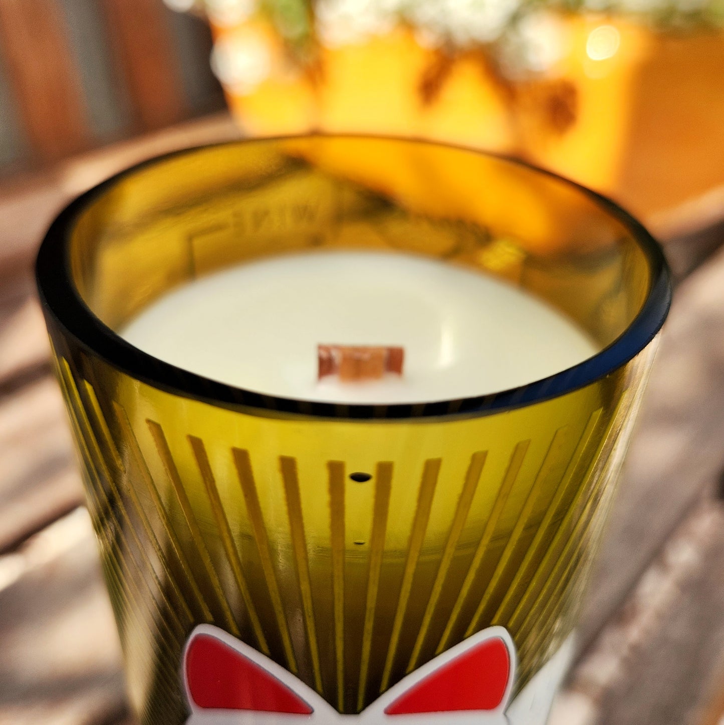 Close-up of a sustainable wooden wick candle housed in a green upcycled bottle featuring red and white label accents. Clean-burning coconut soy wax is poured into a smooth glass vessel with a polished rim for an artisanal touch.