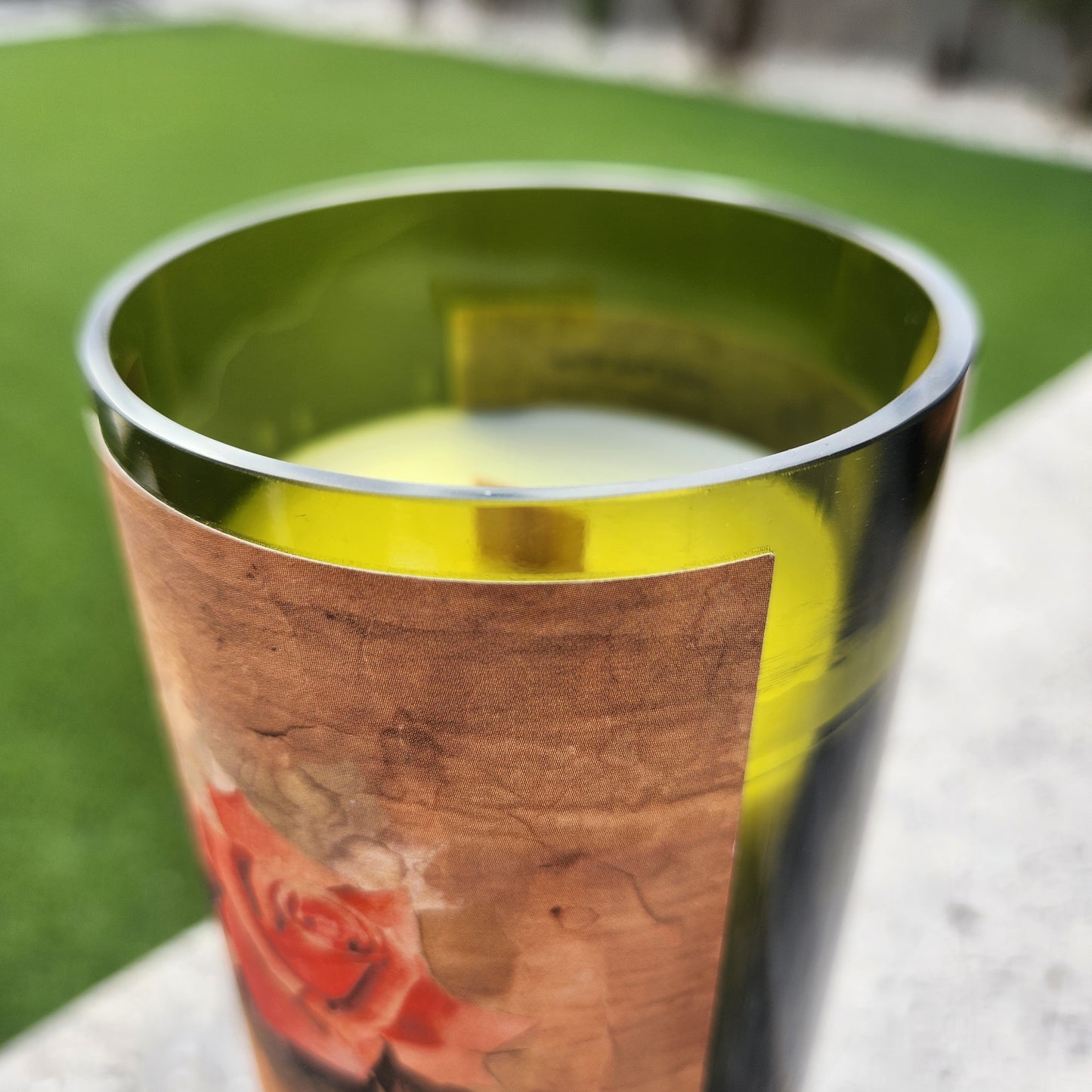 Close-up of the rim of a recycled wine bottle candle made from a Dos Cabezas pink wine bottle, highlighting the glossy smooth edge and pale green tint of the upcycled glass. The candle inside features a clean-burning wooden wick and creamy white coconut soy wax, with a portion of the rustic rose label visible for a handcrafted, vintage-inspired aesthetic.