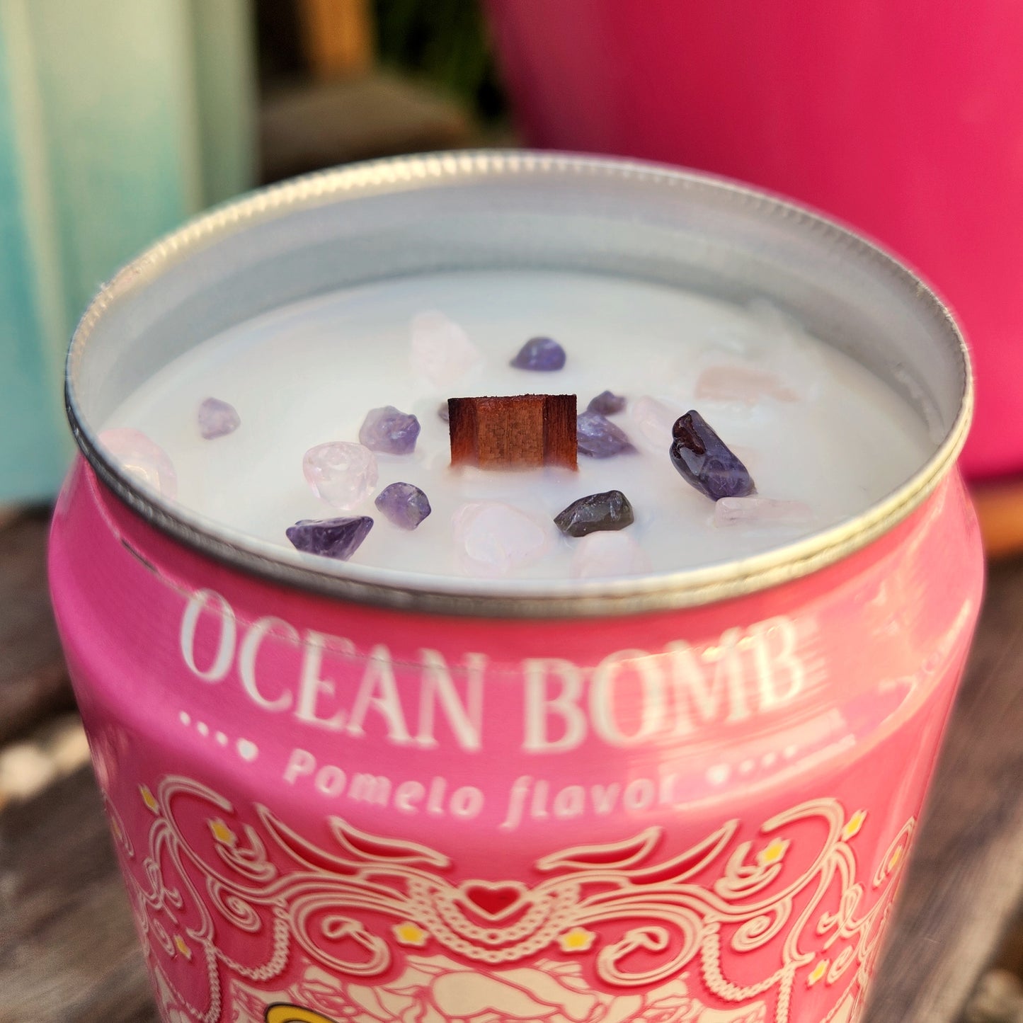 A close-up of the rim of a pink Ocean Bomb can candle, showing a crackling wooden wick and a sprinkle of rose and purple crystals atop smooth white coconut soy wax.