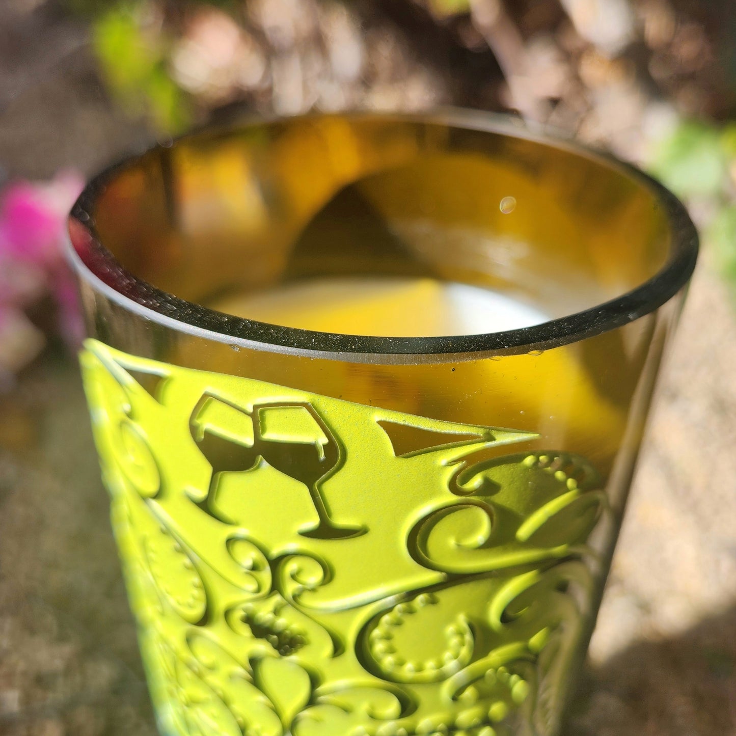 Close-up of a hand-poured, eco-friendly coconut soy wax candle with a wooden wick, set in a recycled wine bottle featuring an embossed skull-and-swirl design; the shiny smooth rim catches sunlight, adding contrast to the textured green label.