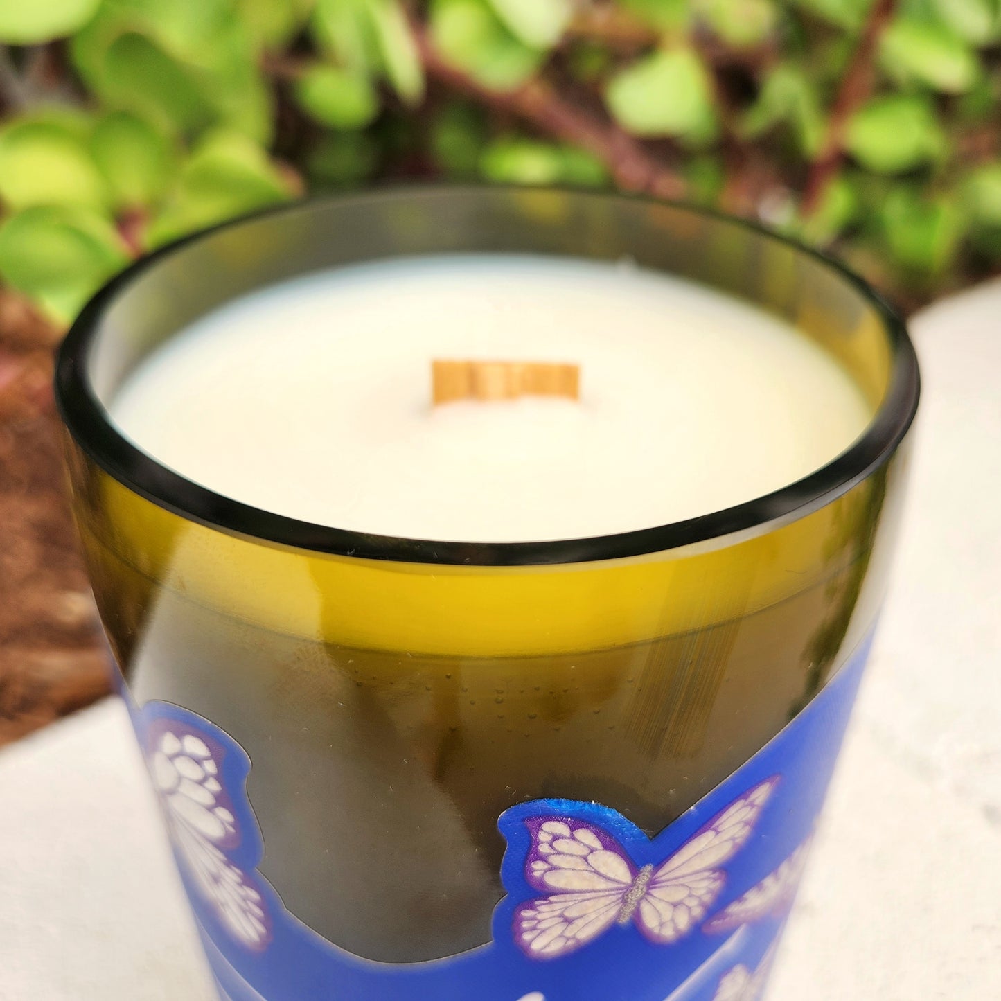 Close-up of a clean-burning candle in a recycled green wine bottle with a smooth glass rim and visible wooden wick; purple butterflies from the Dolly-themed label curve around the side.
