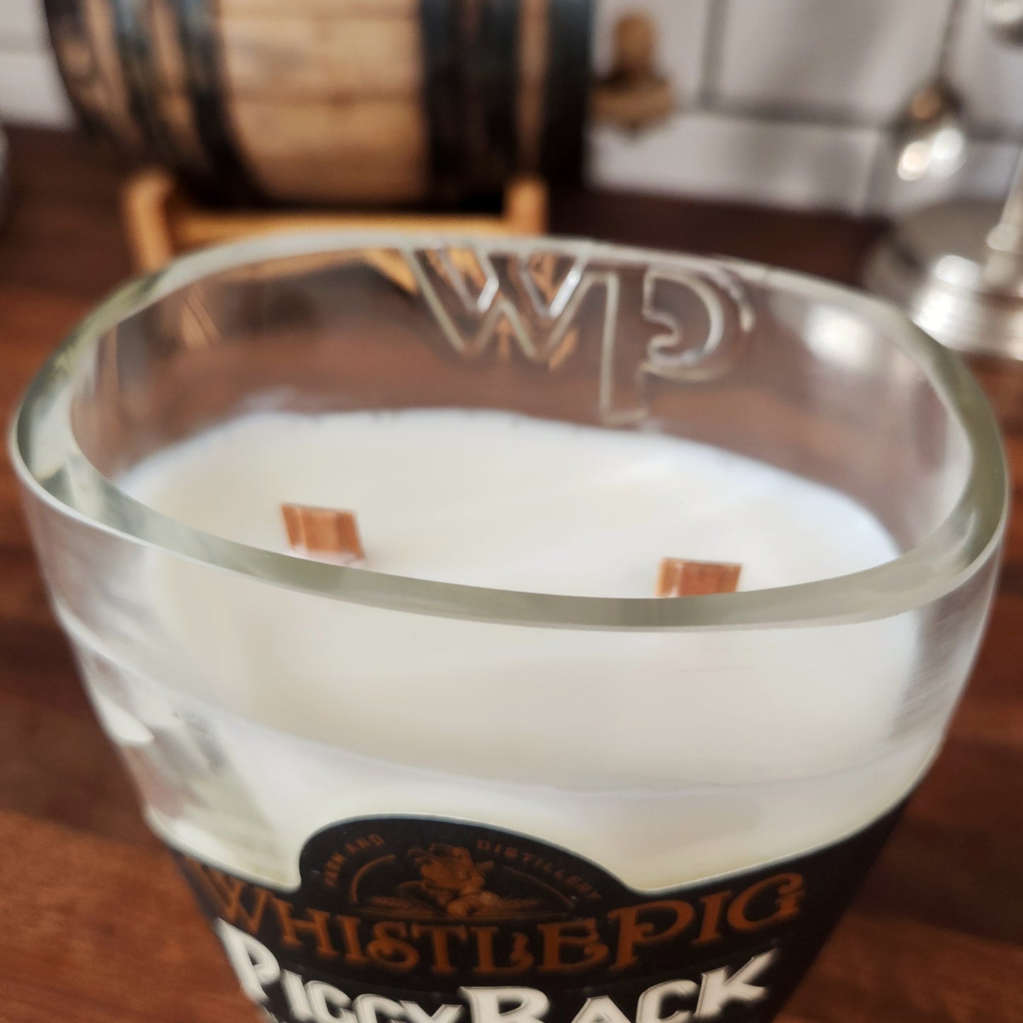Close-up of a clean-burning, non-toxic coconut soy wax candle with two wooden wicks, poured into a uniquely shaped recycled whiskey bottle featuring an embossed “WP” and the WhistlePig PiggyBack label on the front.