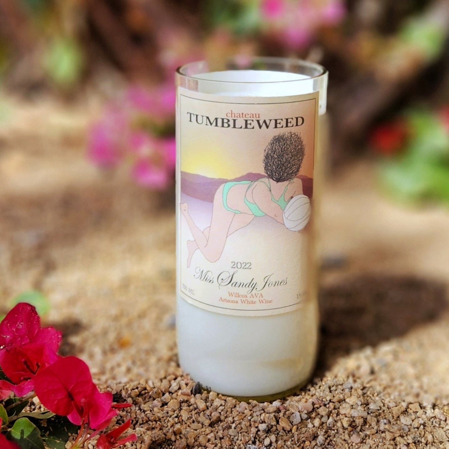 A hand-poured coconut soy wine bottle candle crafted from a recycled Chateau Tumbleweed Miss Sandy Jones bottle. The label features a playful beach volleyball illustration set against a warm desert sunset. The eco-friendly wooden wick candle has a smooth, polished glass rim, showcasing expert craftsmanship. Placed in a natural desert setting with vibrant pink bougainvillea flowers and sandy earth, this sustainable wine bottle candle is a perfect blend of Arizona charm and artisan luxury.