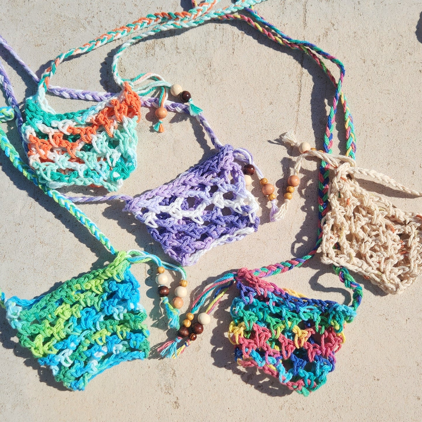 A colorful set of five hand-crocheted wine glass lanyards, each featuring a unique pattern and color palette. Made with braided cotton yarn and finished with wooden bead accents, the lanyards are laid out on a sunlit concrete surface, showcasing their playful textures and festival-ready charm.