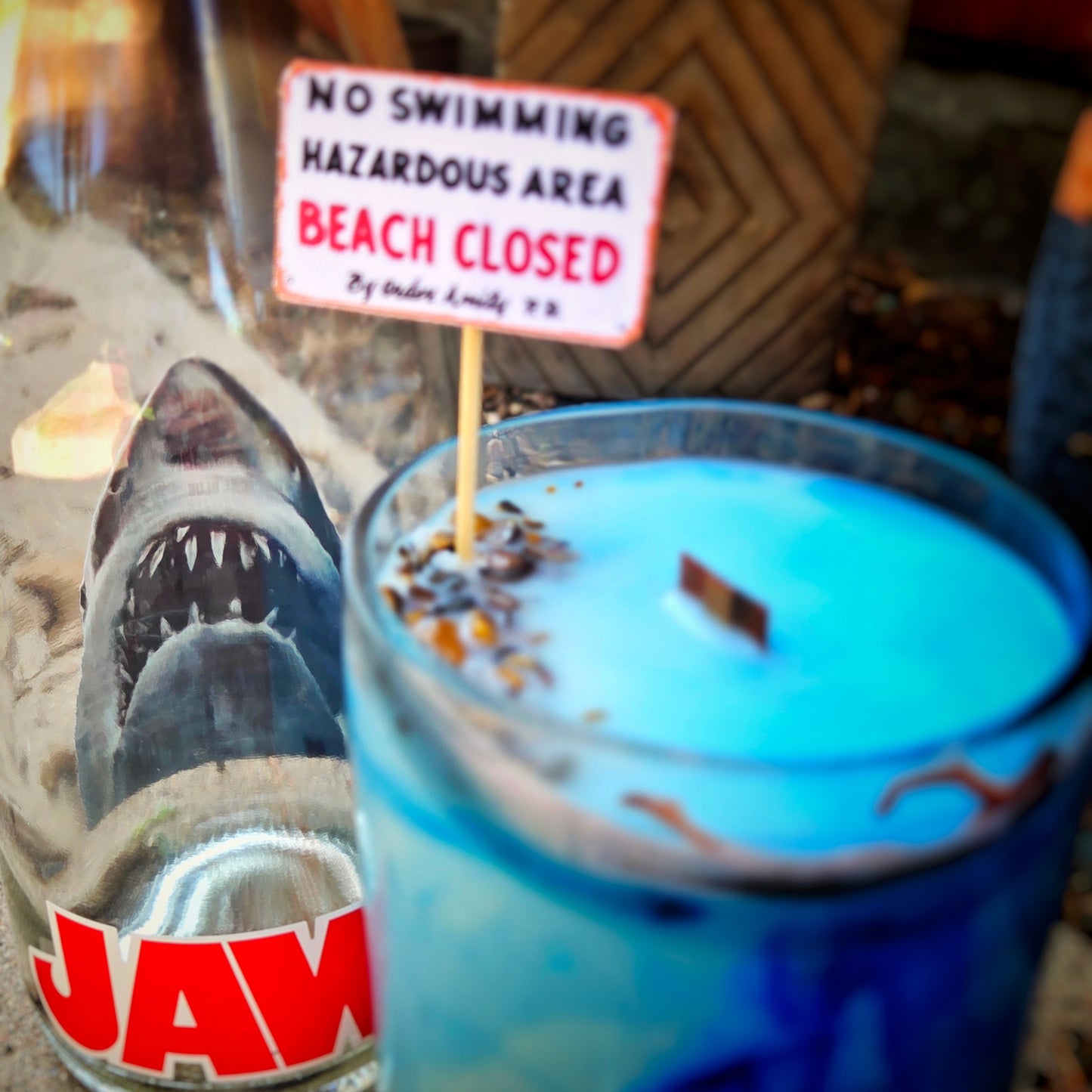 Close-up of a hand-poured wine bottle candle with ocean-blue wax and tiger's eye crystals, positioned in front of a shark face graphic, highlighting the âNo Swimmingâ beach closure sign and wooden wick.