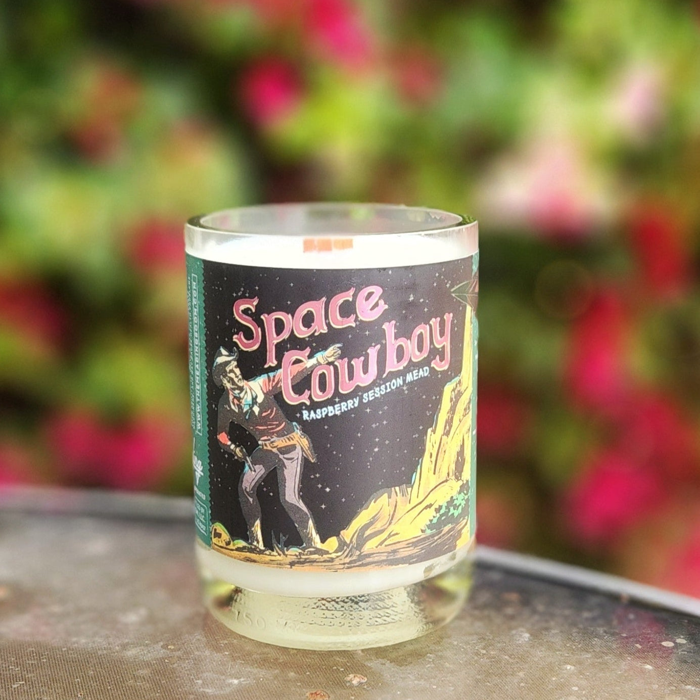 A hand-poured coconut soy wax candle crafted from a recycled Space Cowboy Raspberry Session Mead bottle. The label features a retro sci-fi cowboy illustration with a cosmic western theme. This eco-friendly wooden wick candle has a smooth, polished glass rim, highlighting expert craftsmanship. Placed on a reflective glass table with a blurred background of lush greenery and pink flowers, this upcycled mead bottle candle is perfect for sci-fi lovers, space western fans, and eco-conscious shoppers.
