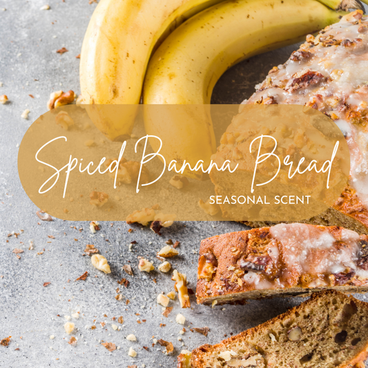 A loaf of spiced banana bread topped with icing and chopped nuts sits next to a bunch of ripe bananas, with scattered crumbs and walnuts on a gray surface, illustrating the warm and cozy Spiced Banana Bread seasonal scent.