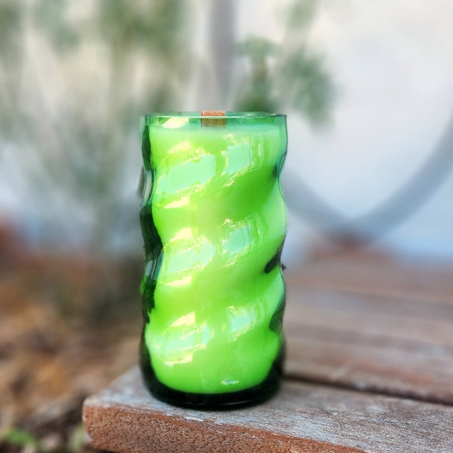 A hand-poured coconut soy wax candle housed in a recycled Squirt soda bottle with a distinctive twisted glass design. The vibrant green hue of the vessel enhances its bold, eye-catching look. This eco-friendly wooden wick candle has a polished glass rim, showcasing expert craftsmanship. Placed on a rustic wooden surface with a softly blurred natural background, this sustainable upcycled soda bottle candle is a unique blend of creativity, sustainability, and nostalgic charm.
