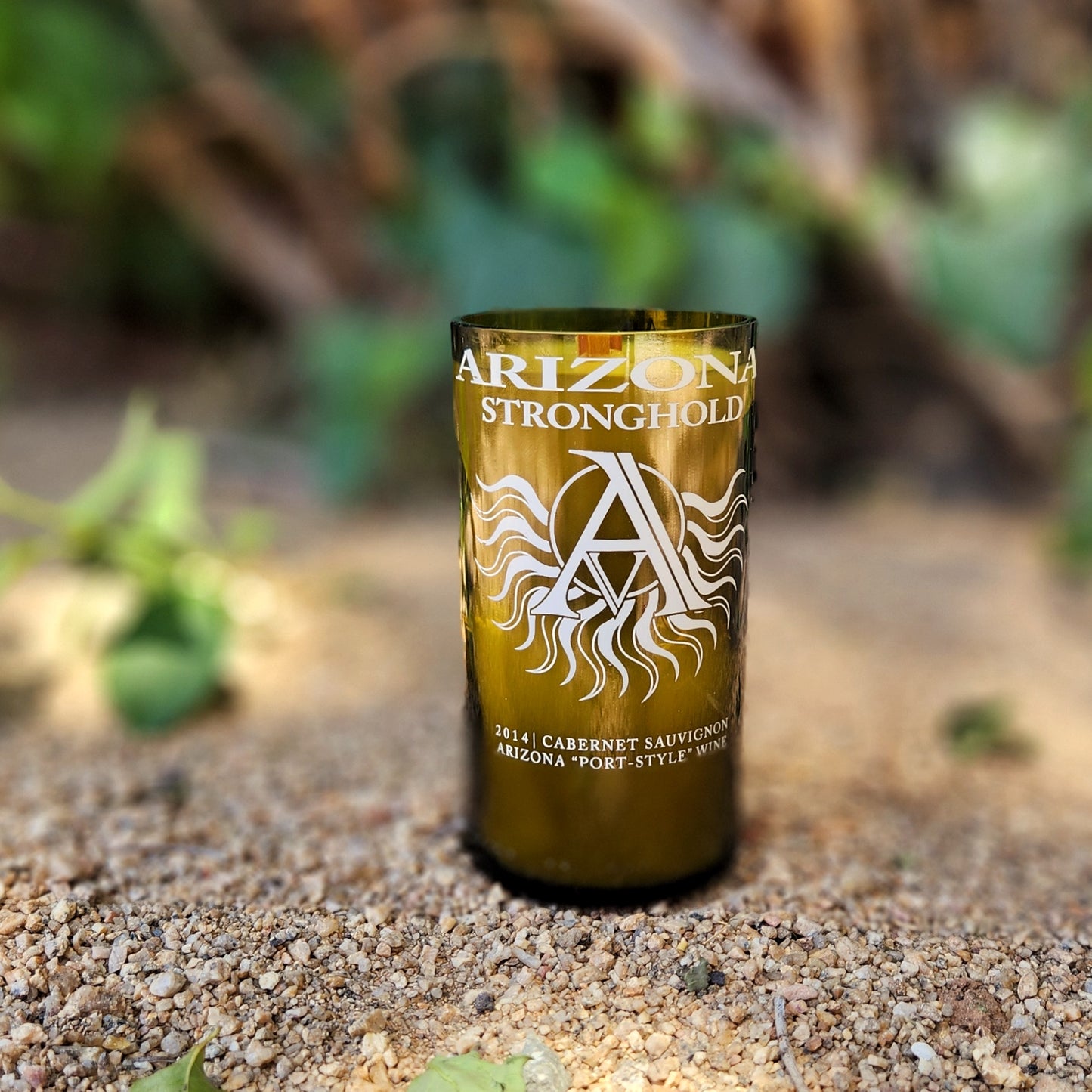 Eco-friendly wine bottle candle made from a recycled Arizona Stronghold bottle, featuring an etched white sunburst logo and bold lettering. The vessel sits upright on sandy ground with soft greenery in the background, emphasizing its natural, clean-burning appeal.