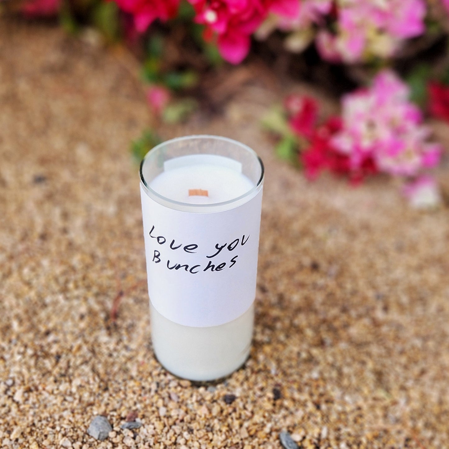 A full-view photo of the “Love You Bunches” candle, made from a recycled wine bottle. Set against a soft desert background with bougainvillea blooms, this wooden wick candle captures charm and sustainability in one cozy, clean-burning design.
