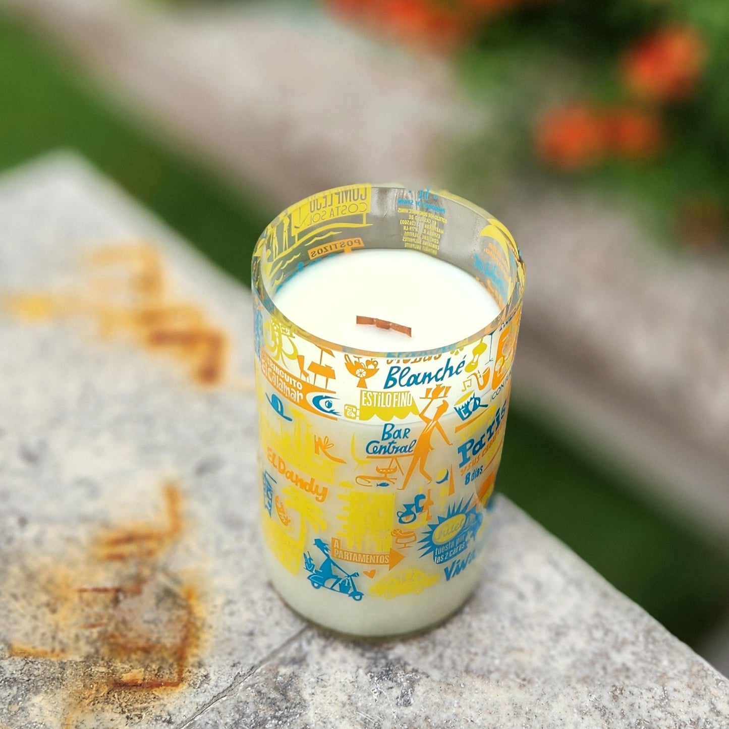 Angled view of the upcycled wine bottle candle, highlighting the clear, glossy rim and vibrant, overlapping label art in orange, yellow, and blue with travel motifs.