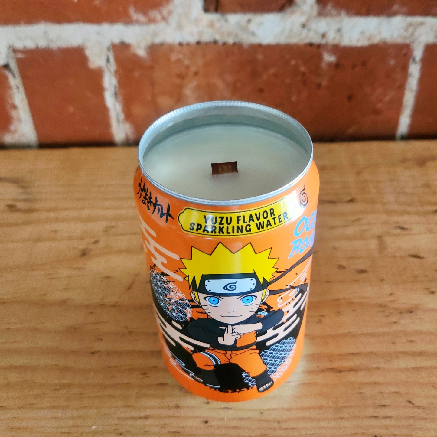 A hand-poured coconut soy candle with a wooden wick inside a vibrant orange recycled can, decorated with a stylized anime character in action pose.