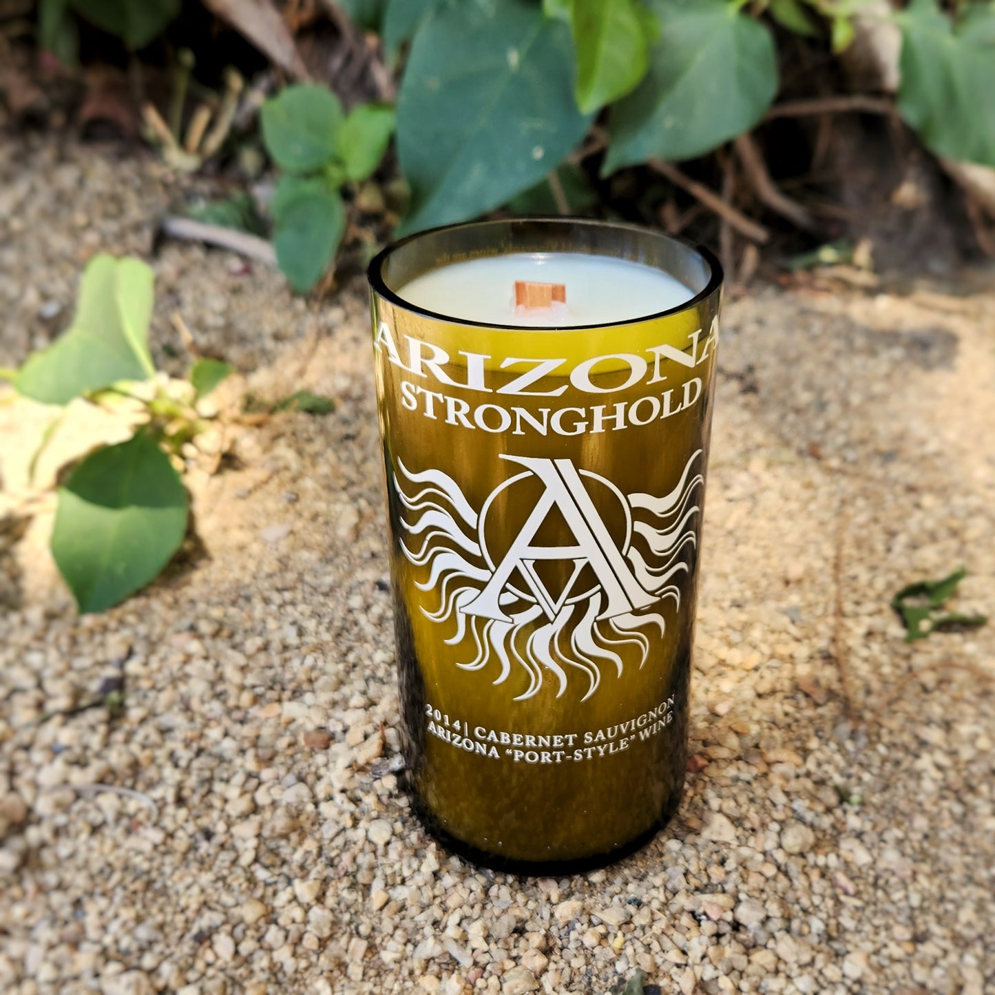 Hand-poured coconut soy wine bottle candle in an upcycled Arizona Stronghold bottle, showcasing a smooth, shiny rim and centered wooden wick. The white etched logo contrasts against the green glass, with a sandy outdoor setting reinforcing its sustainable charm.