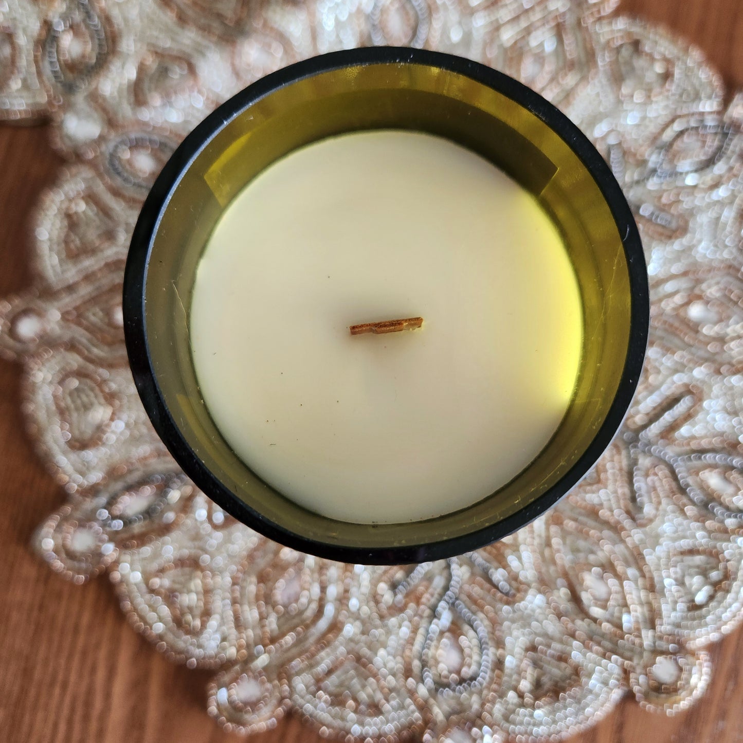 Top-down view of a hand-poured coconut soy wax candle with a wooden wick, housed in a smooth-rimmed recycled wine bottle. The glossy wax surface sits clean and even inside deep green glass, resting on a detailed beaded placemat. Eco-friendly, non-toxic burn.