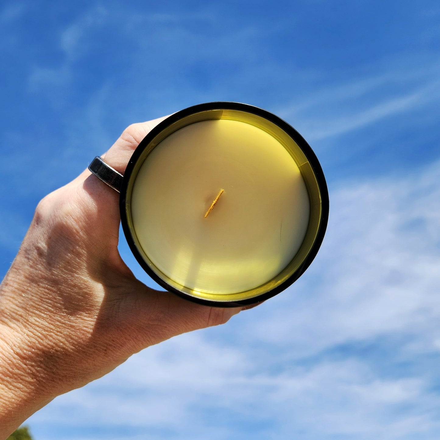 A top-down view of a hand-poured Fusion wine bottle candle with a wooden wick, held up against a vivid blue sky. The translucent amber glass rim showcases the eco-friendly upcycled wine bottle vessel, filled with creamy coconut soy wax.