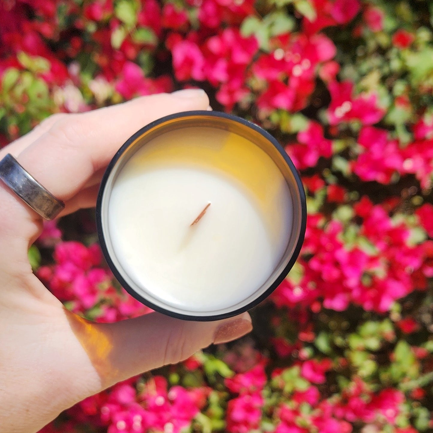 Top-down view of an eco-friendly candle poured in a recycled amber soda bottle. The creamy coconut soy wax is centered by a wooden wick, glowing against a backdrop of bright pink blossoms for a bold, festive vibe.