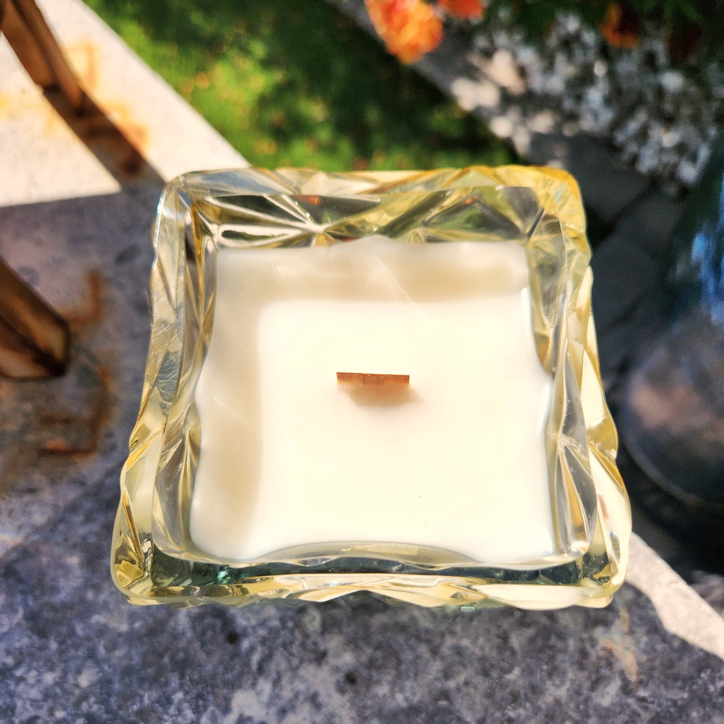 Top view of a square-shaped Mi Familia Flores bottle candle with smooth coconut soy wax and a centered wooden wick, set in textured, light-catching glass.