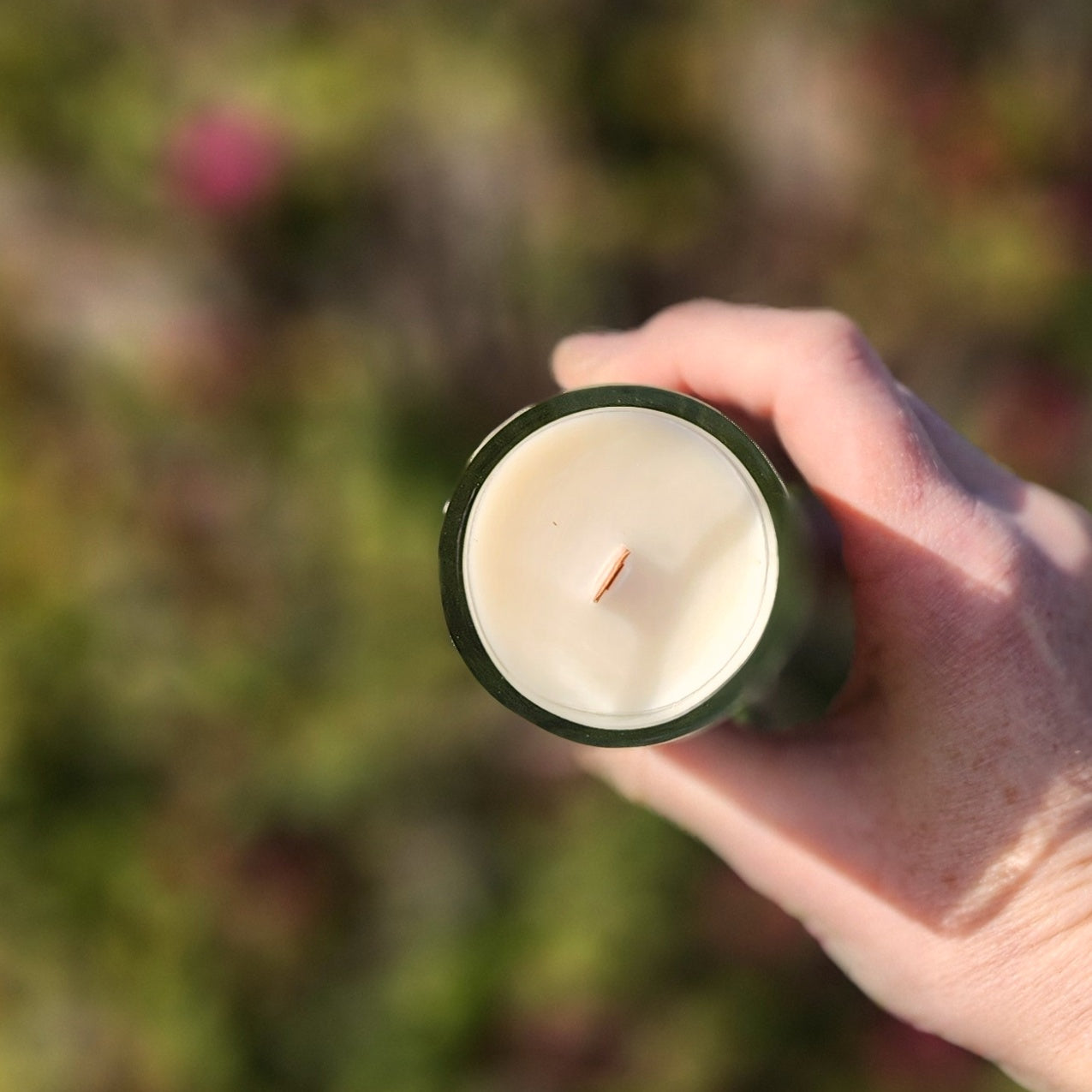 Top-down view of a hand holding a white coconut soy wax candle with a centered wooden wick, set in a recycled wine bottle with a polished glass rim. Captures the eco-friendly craftsmanship of this clean-burning Rune Mead candle against a blurred green garden backdrop.