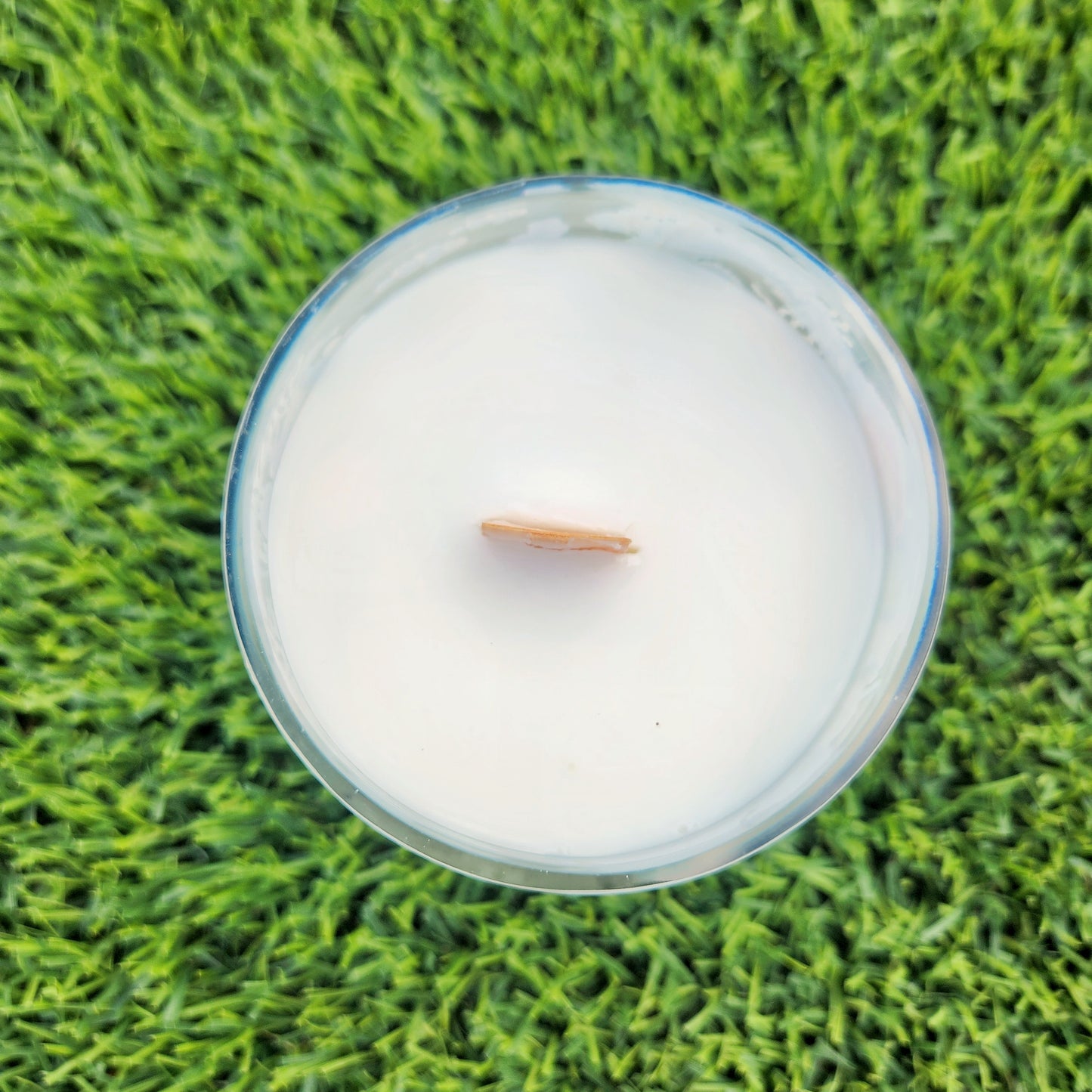 Top-down view of a hand-poured candle in a clear wine bottle with a smooth, polished rim, filled with creamy white coconut soy wax and centered with a wooden wick. Set against vibrant green grass, this clean-burning candle highlights eco-friendly elegance.