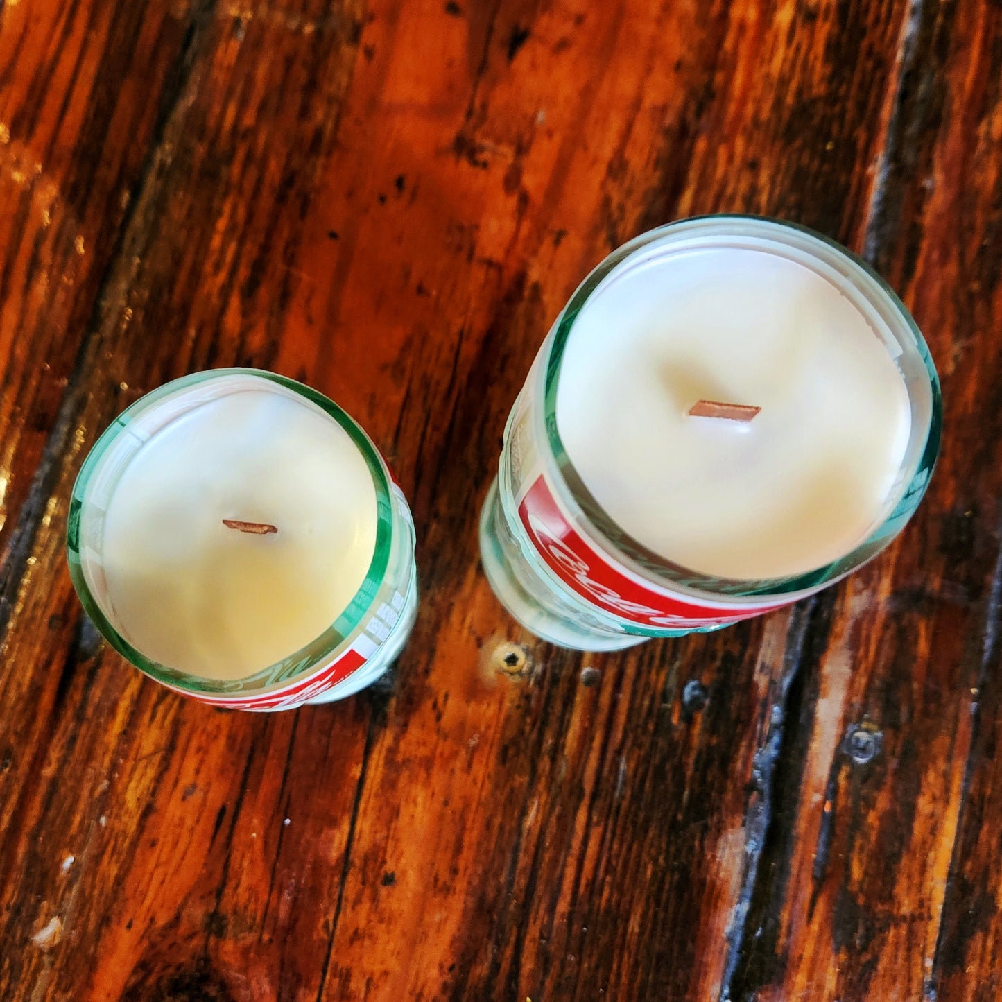 Top-down view of two eco-friendly coconut soy candles with wooden wicks, hand-poured into recycled Coca-Cola glass bottles, shown on a rustic wood table.