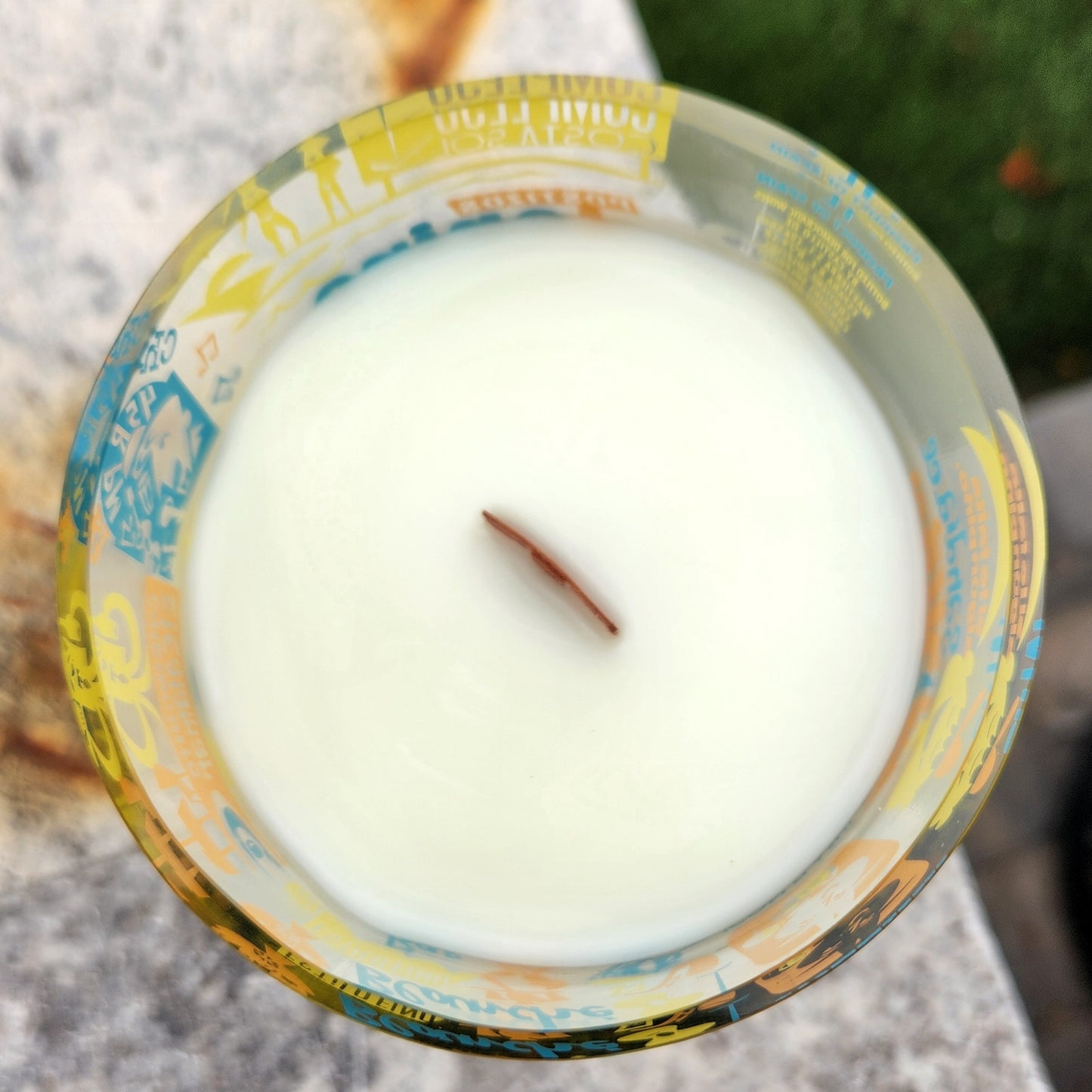 Top-down view of an unlit eco-friendly wine bottle candle showing creamy coconut soy wax and a centered wooden wick; the surrounding glass is covered in bold, vintage-style illustrations.