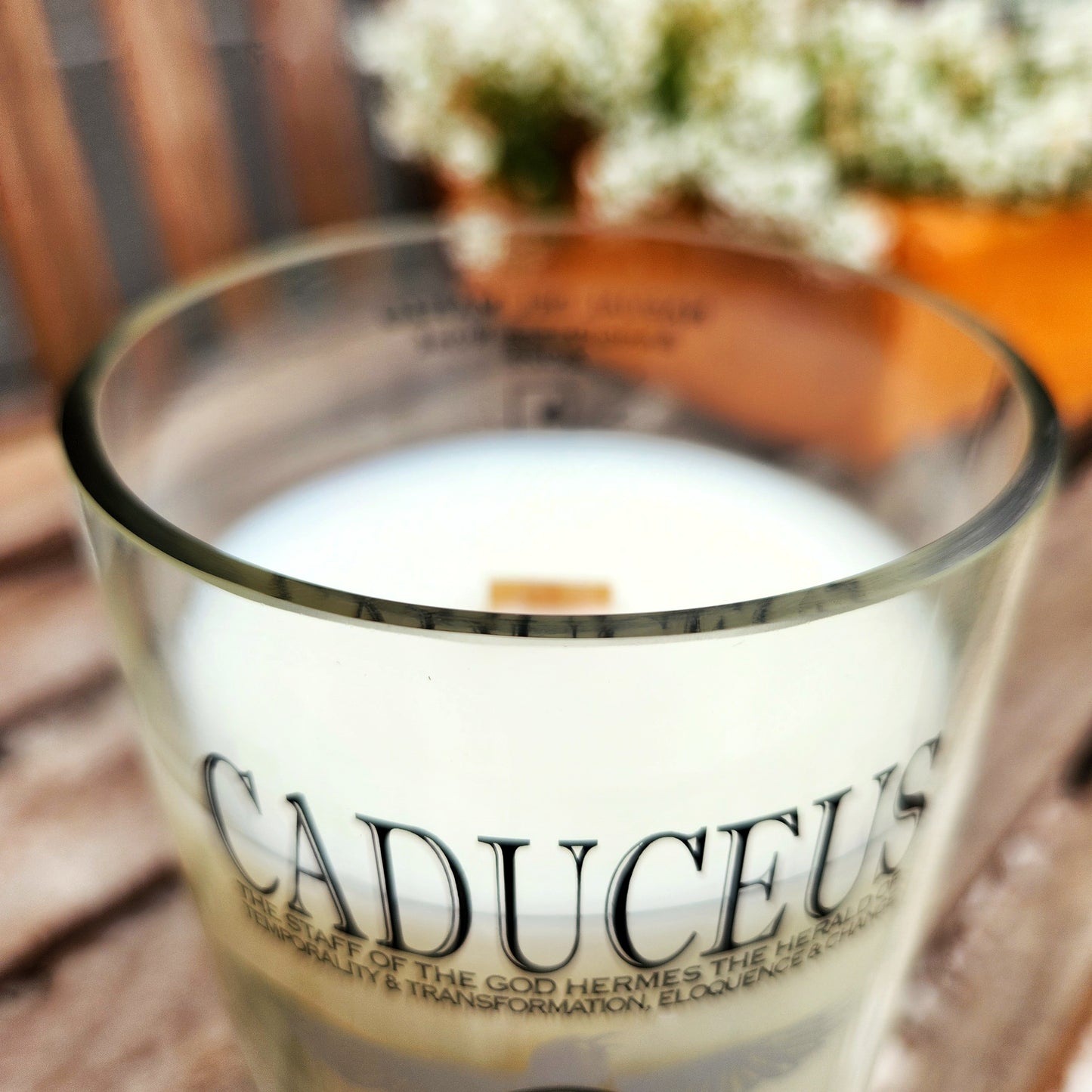 A close-up rim view of a recycled Caduceus wine bottle candle, showing the smooth, glossy glass edge and the wooden wick centered in creamy white coconut soy wax. The bold "Caduceus" logo wraps around the glass, with crisp black lettering that pops against the clean wax. Eco-friendly and hand poured, this wine bottle candle sits on a rustic wooden bench with blurred flowers in the background.