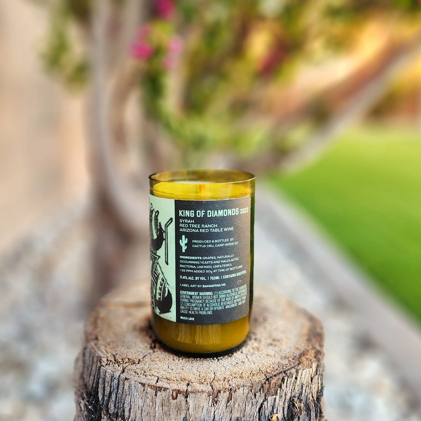 A hand-poured wine bottle candle displayed on a tree stump outdoors, featuring a sharp-cut rim and bold black label reading “King of Diamonds.” The eco-friendly vessel is filled with smooth coconut soy wax and topped with a wooden wick.
