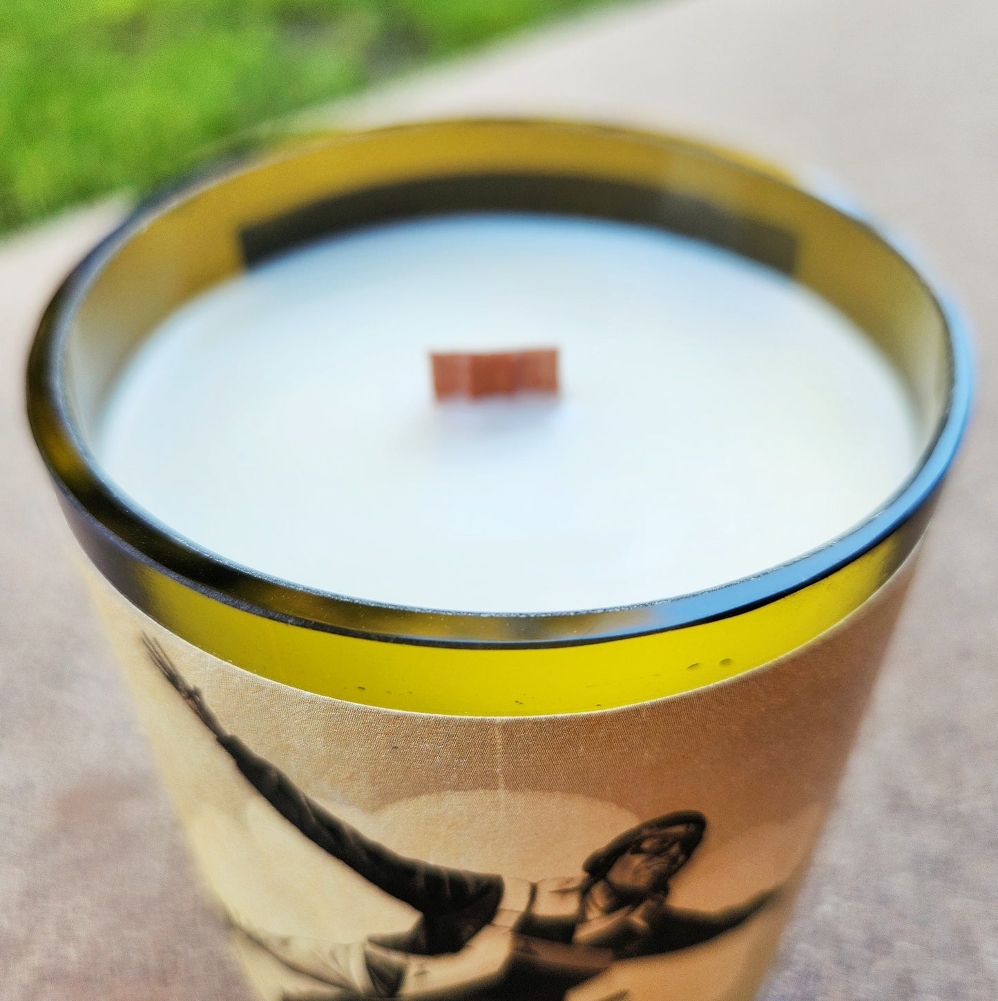Close-up of a hand-poured, eco-friendly wine bottle candle with a wooden wick and a shiny smooth rim. The creamy coconut soy wax is clean and unlit, and the repurposed wine bottle label features a vintage bomber-style graphic with a skydiver.
