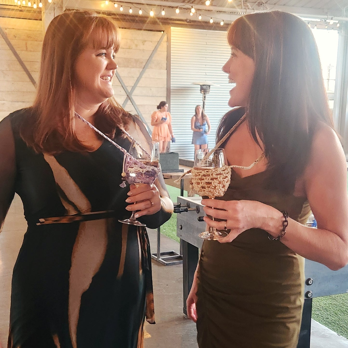 Two women smiling and chatting at an indoor event while wearing handmade crochet wine glass lanyards around their necks, each holding a stemmed glass nestled securely in a knit holder—one in purple and the other in a cream-colored design. String lights glow overhead, with people mingling in the softly lit background.