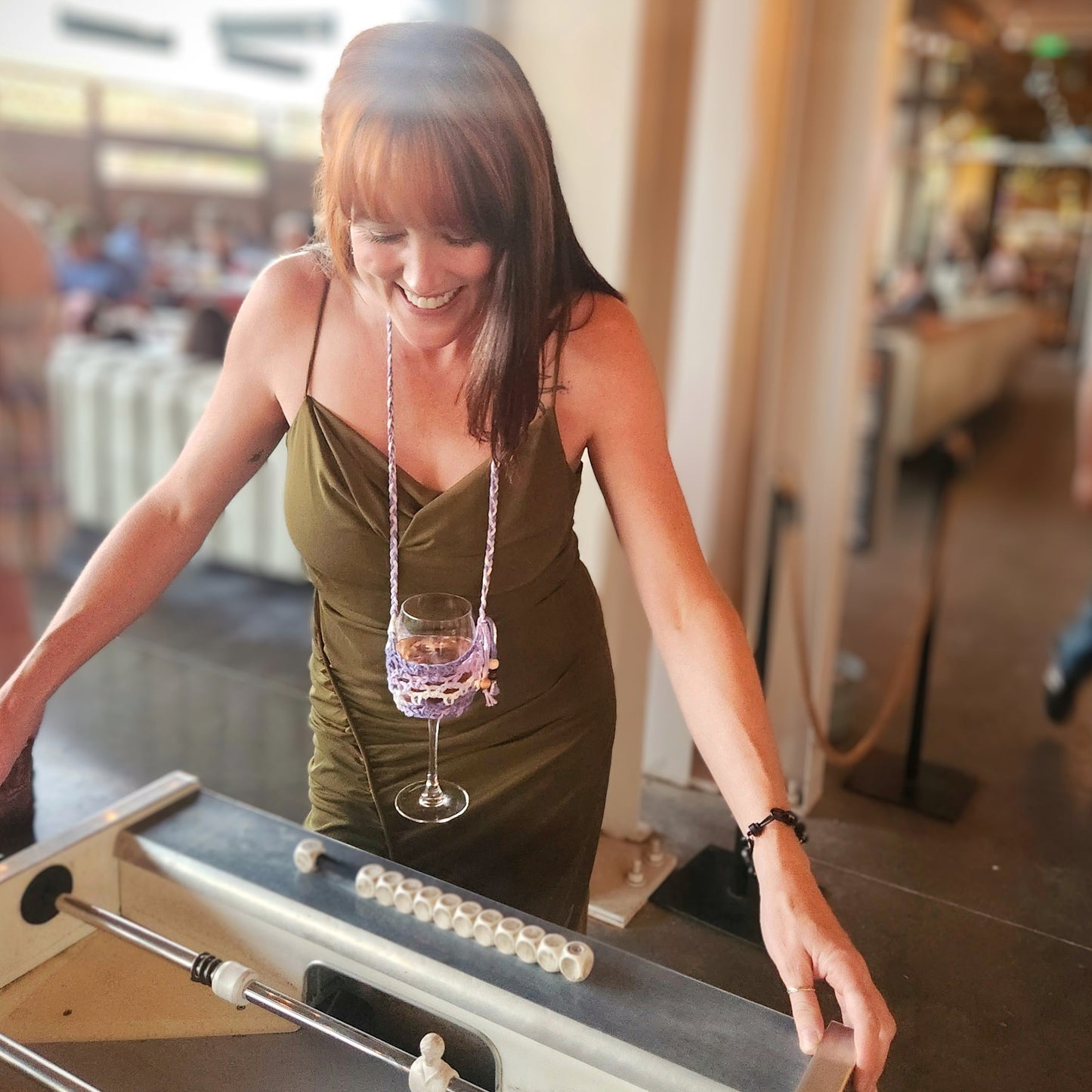 A smiling woman in a green dress plays foosball while wearing a handmade crochet lanyard that holds a wine glass at chest height. The lanyard is crocheted in shades of purple and white, allowing her to enjoy her drink hands-free. The setting is a lively indoor venue with a blurred background of guests and cozy seating, capturing a playful and social atmosphere.
