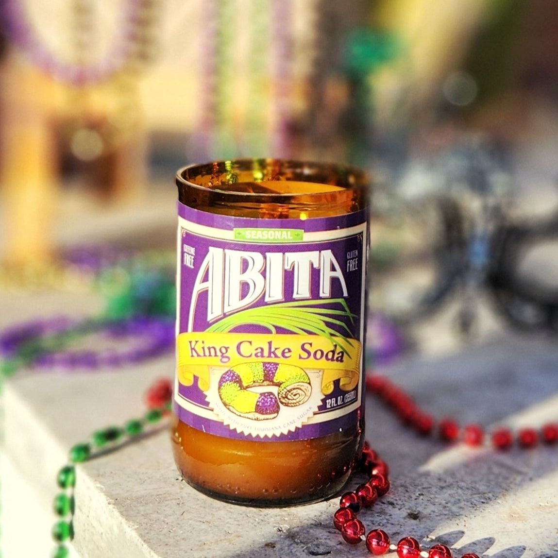A hand-poured coconut soy wax candle made from a recycled Abita King Cake Soda bottle. The festive label showcases bright Mardi Gras colors with an illustration of a King Cake, celebrating the spirit of New Orleans. This eco-friendly wooden wick candle has a polished glass rim, highlighting its artisanal craftsmanship. Placed on a sunlit stone surface surrounded by colorful Mardi Gras beads, this upcycled soda bottle candle is perfect for fans of New Orleans culture and sustainable gifts.