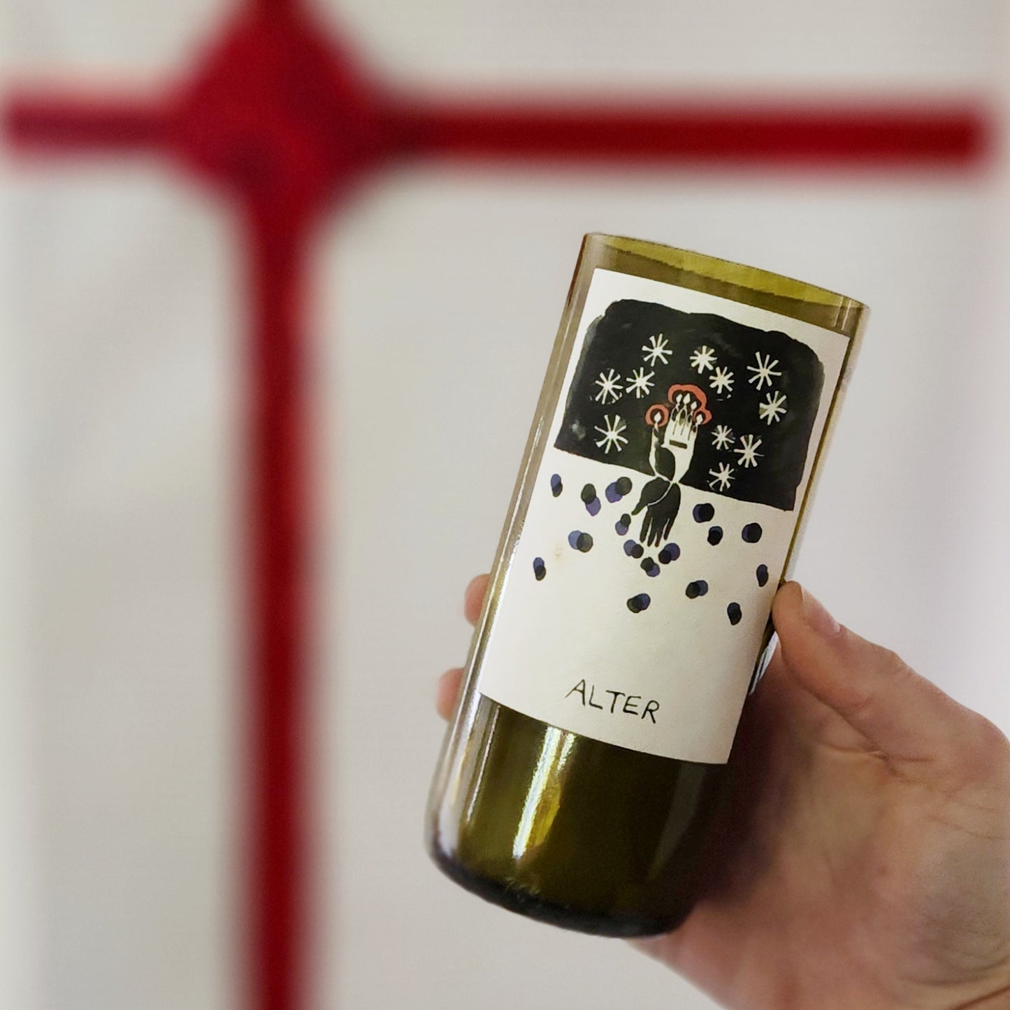 A hand holding a recycled wine bottle candle with a smooth glass rim, featuring a festive label depicting a hand holding candles amidst a starry night. The word "Alter" is printed below the artwork. In the background, a red Christmas bow on a blurred white wall adds a cheerful holiday atmosphere, complementing the seasonal vibe of the candle.