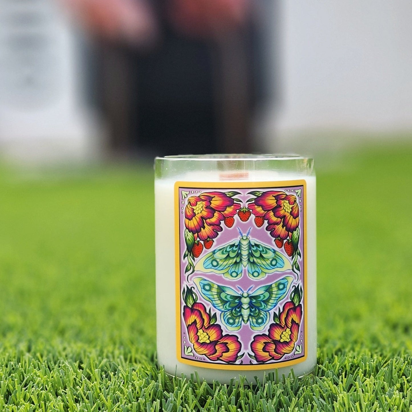 A handcrafted candle housed in a recycled glass mead bottle, featuring a vibrant label with a symmetrical design of a green luna moth surrounded by blooming red and yellow flowers and delicate strawberries. The candle sits on lush green grass, with a blurred background of pink flamingo yard decorations, creating a whimsical and artistic outdoor setting. The smooth glass rim and creamy white wax enhance its artisanal and eco-friendly appeal.