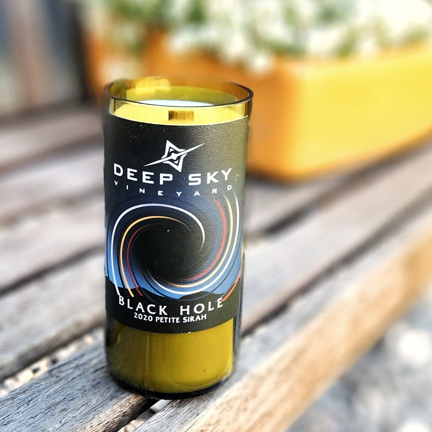 A recycled glass candle crafted from a green wine bottle, featuring a sleek black label from Deep Sky Vineyard. The label displays a swirling, cosmic-inspired design with the text "Black Hole 2020 Petite Sirah." The candle is placed on a wooden bench outdoors, with vibrant planters filled with greenery and flowers in the blurred background, creating a fresh and inviting atmosphere. The wooden wick and clean finish of the glass rim add a touch of artisanal elegance.