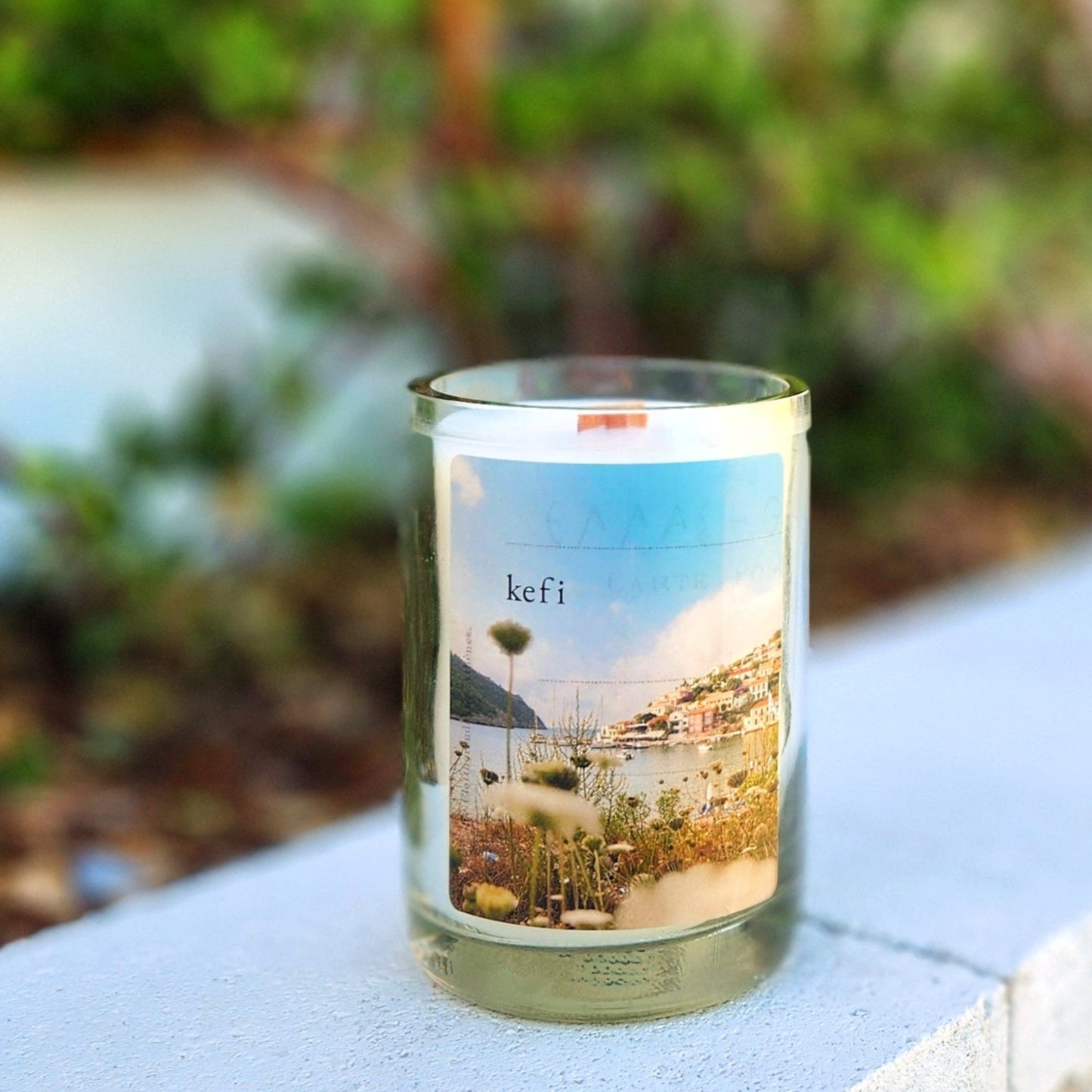 A handcrafted candle housed in a recycled glass mead bottle, featuring a serene label with a picturesque coastal scene, including a harbor, colorful hillside buildings, and wildflowers in the foreground. The label is titled "kefi," evoking a sense of joy and inspiration. The candle is set outdoors on a light concrete surface, with a blurred backdrop of greenery and sunlight, enhancing its tranquil and eco-friendly charm. The smooth glass rim and wooden wick add a touch of artisanal elegance.