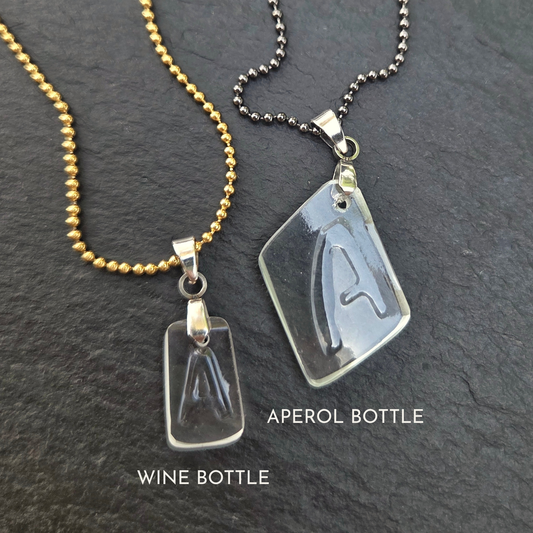 Two handmade Letter A necklaces made from reclaimed bottle glass, showing the smaller Wine Bottle pendant on a gold-tone chain and larger Aperol Bottle pendant on a gunmetal chain.