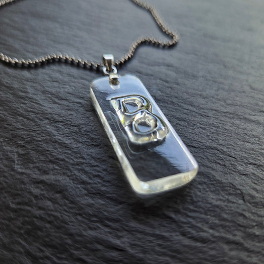Angled close-up of a clear reclaimed whiskey bottle glass Letter B pendant showing the original molded lettering, three-dimensional raised surface, and thick polished glass edges on dark slate.